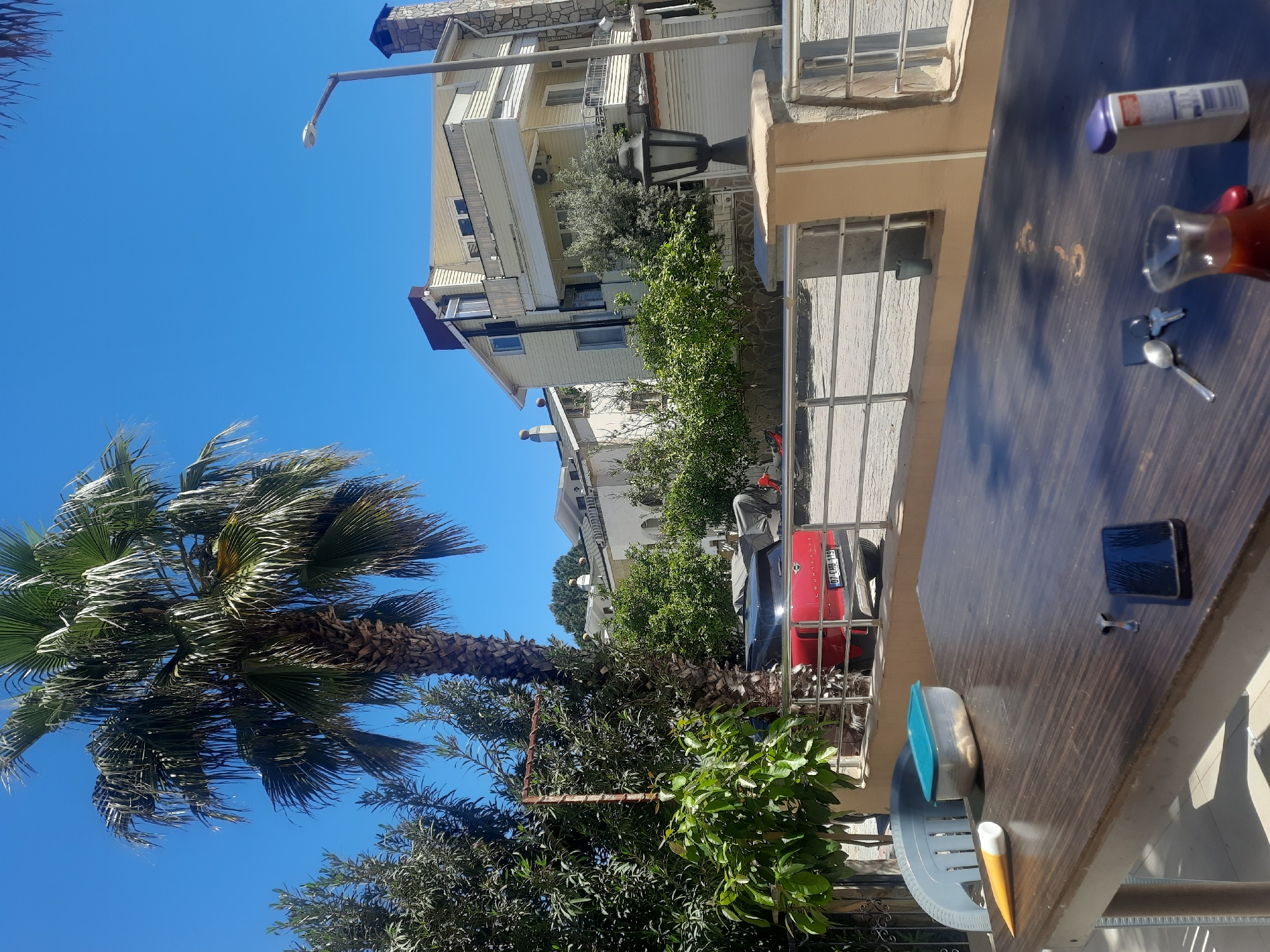 The image captures a scene in Kemer, Turkey, under a clear blue sky. A tall palm tree stands prominently in the left side of the frame, its fronds extending towards the sky. A building with multiple levels, painted in light colors, is visible behind the trees.
In the foreground, there's a wooden table set outdoors, with personal items scattered on its surface, including a smartphone, a small glass cup with tea, keys, a plastic food container, and a bottle of cream. A pen lies on the table edge. A red car is parked behind the table. The photo appears to have been taken at daytime in a residential area.