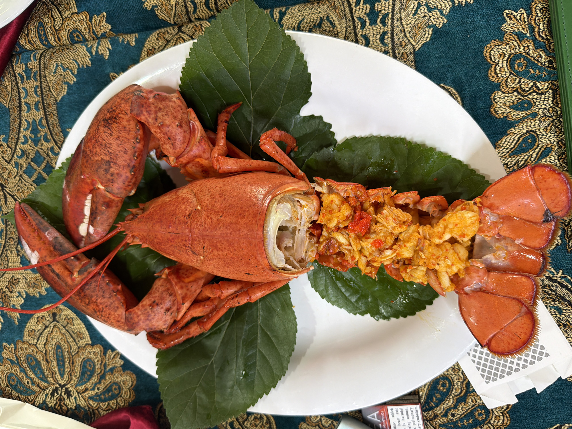 A cooked whole lobster, presented on a white plate with green leaves, sits on a table covered with a teal and gold patterned tablecloth. The lobster is bright red and has its tail and claws separated from the body. The body cavity is filled with a mixture of shredded lobster meat and some red roe. The background shows a close-up of the ornate tablecloth and the edge of a white napkin with a packet of "LA Menthol" cigarettes. The scene is lit from above. The image was taken in Kemer, Turkey.