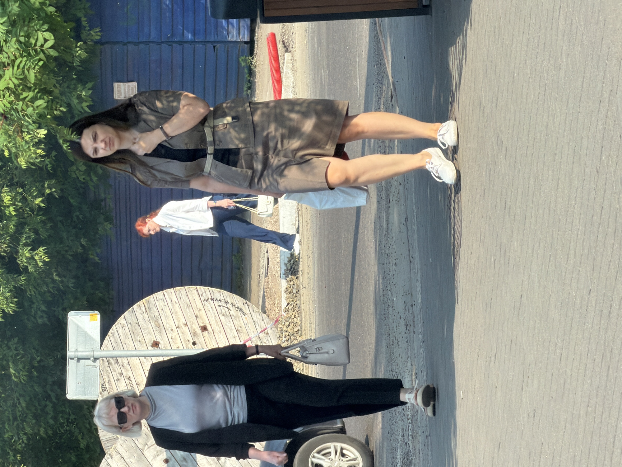 In Krasnoyarsk, Russia, on a sunny day, three women are walking on a paved surface adjacent to a road. The woman in the foreground on the right is wearing a brown suit with a black top and white sneakers. She is carrying a white bag in her left hand and has her right hand on her chin. To her left, another woman with blonde hair and sunglasses is wearing a black jacket over a grey top and black pants, carrying a beige handbag. In the background, a third woman with red hair is wearing a white shirt and blue pants, holding a white bag. The environment features large wooden spools, a blue fence, and trees.