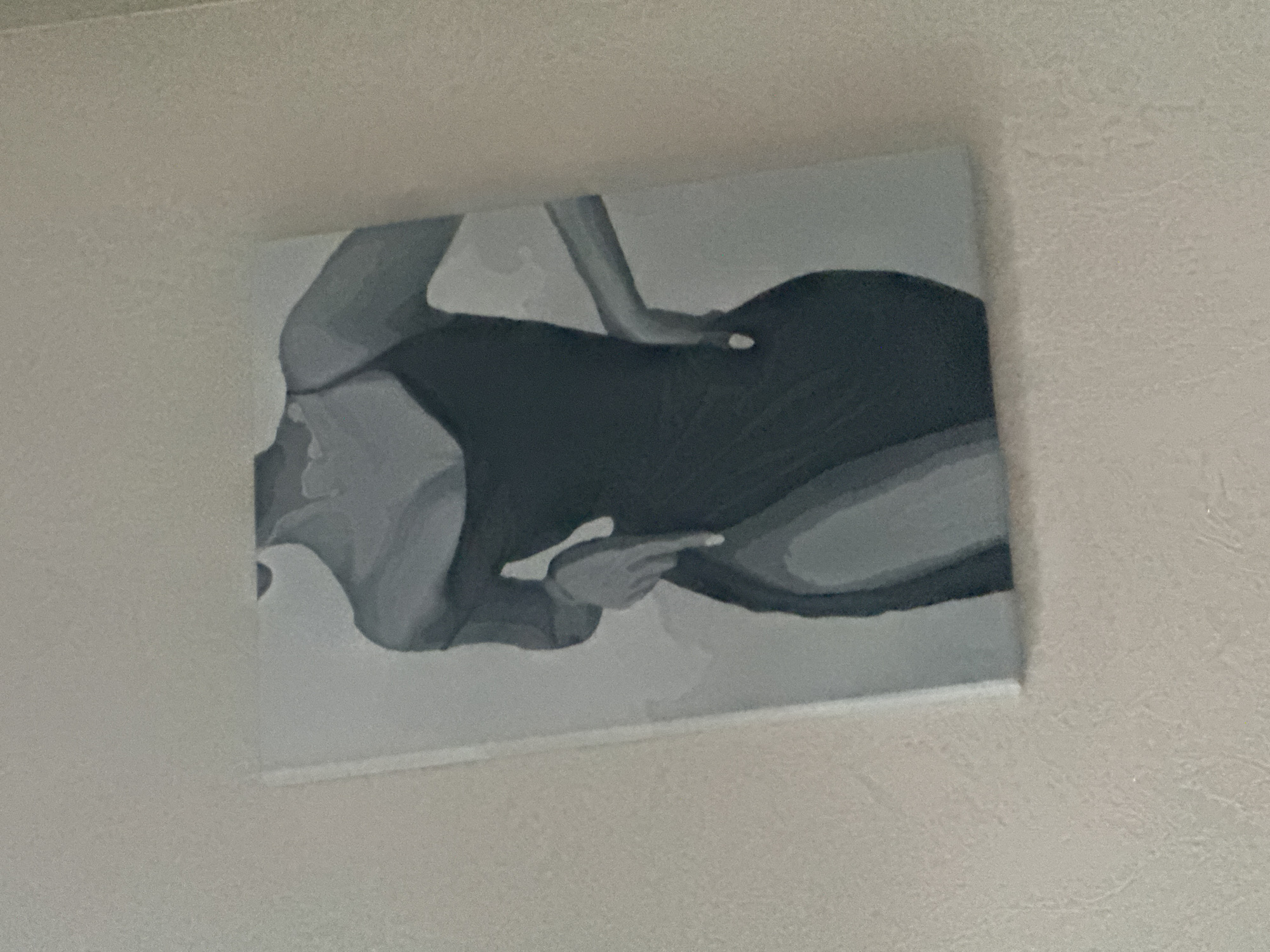 The media file shows a grayscale painting of a person in a reclining or partially seated position, with their body angled towards the left side of the canvas. The painting is abstract, utilizing broad strokes and varying shades of gray to depict the form and musculature of the figure. The subject appears to be wearing dark clothing, possibly a swimsuit or leotard, which contrasts with the lighter tones used for the skin and background. One arm is bent and raised, with the hand visible near the head or shoulder area. The pose suggests a moment of relaxation or contemplation. The painting is mounted on a white wall, which has a textured surface that appears slightly uneven. The lighting suggests an indoor setting, possibly a room with ambient light. There are no visible text elements, and no specific time of day, weather conditions, or strong emotional cues can be definitively inferred from the abstract nature of the artwork and the overall composition. The context provided indicates the location is Saratov, Russia.
