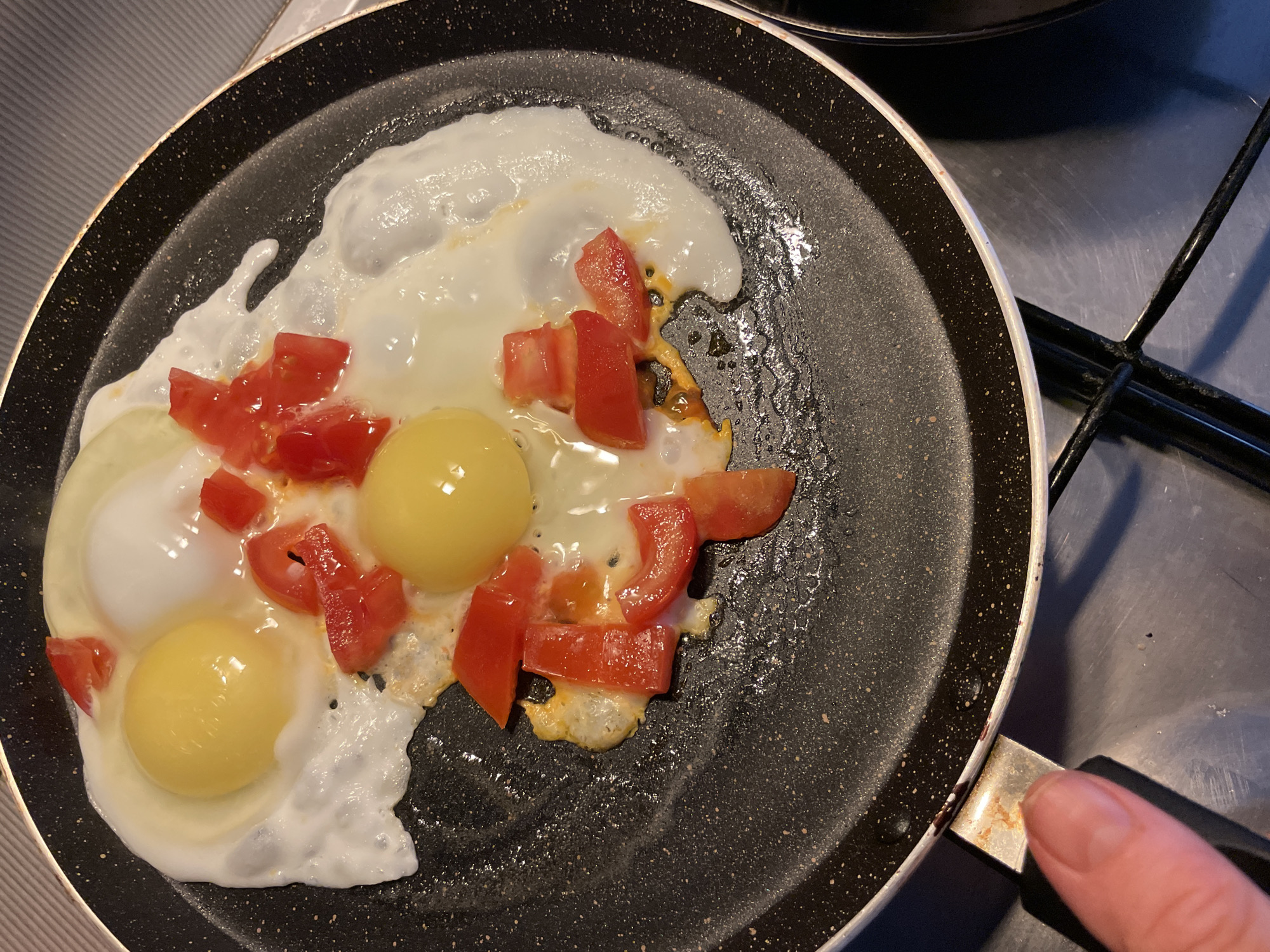 A close-up, top-down shot shows two fried eggs with their yolks intact, interspersed with chopped red tomatoes, cooking in a black non-stick frying pan. The pan is positioned on a stainless steel stovetop, which is part of a kitchen environment. A human finger is visible in the lower left corner, appearing to touch the handle of the frying pan. The image focuses on the food preparation activity. There is no identifiable information regarding the city of Sochi, Russia, or any other specific location.