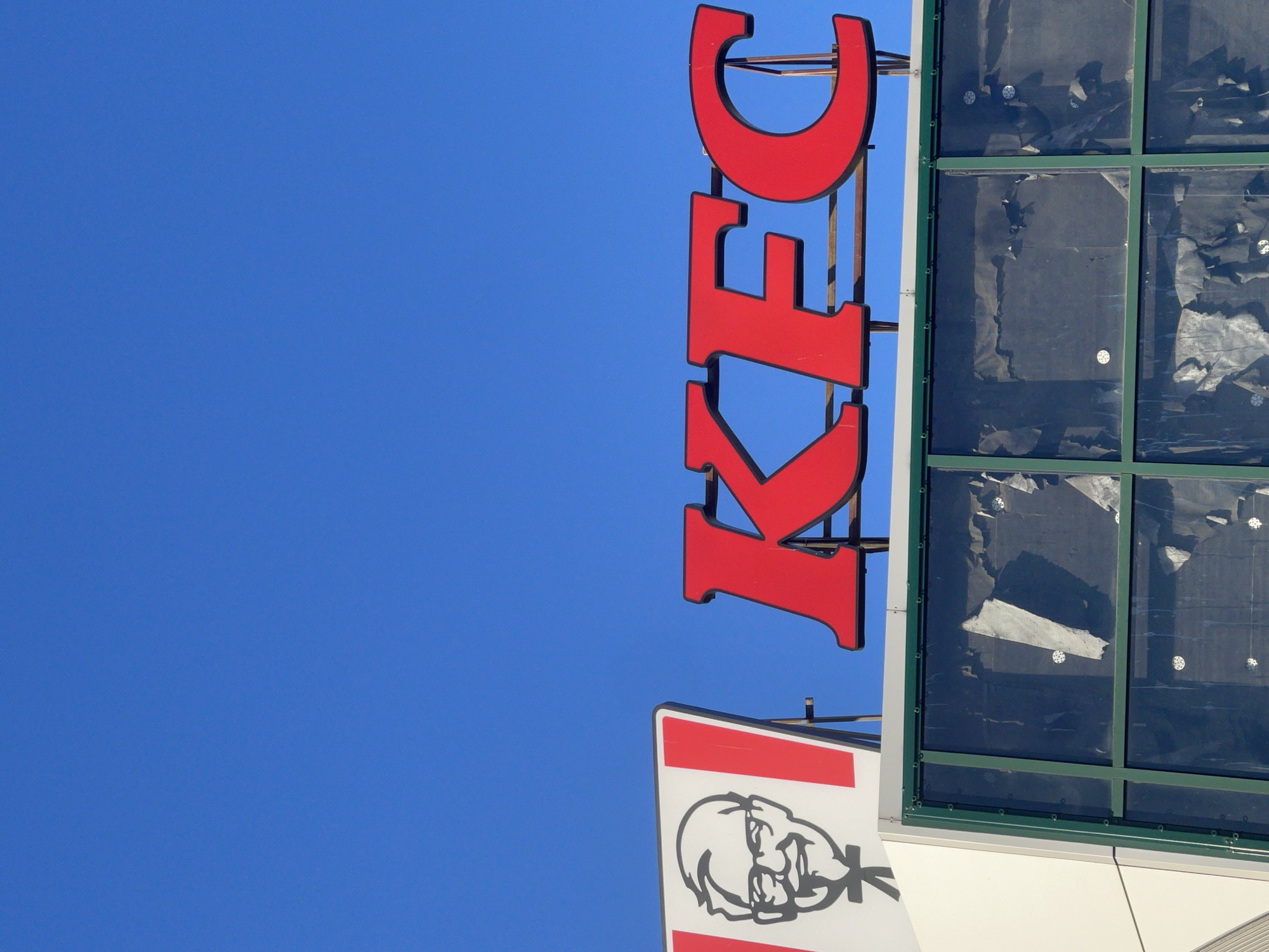 This media file captures a close-up view of a KFC restaurant's signage against a clear blue sky. The primary subject is the iconic red "KFC" logo, rendered in large, bold, three-dimensional letters, oriented vertically. To the left of the letters, a portion of a white sign featuring a black and white illustration of Colonel Sanders is visible. To the right of the KFC logo, the building's facade is present, with large windows covered by dark, opaque material. The windows are framed in green, and small white circular fasteners are visible through the covering. The overall impression is one of a commercial establishment, likely during daytime given the bright sky. There are no people visible in the frame, and no specific activity or event is depicted. The image is cropped tightly on the signage and a section of the building, with the vast expanse of the sky dominating the left side.
