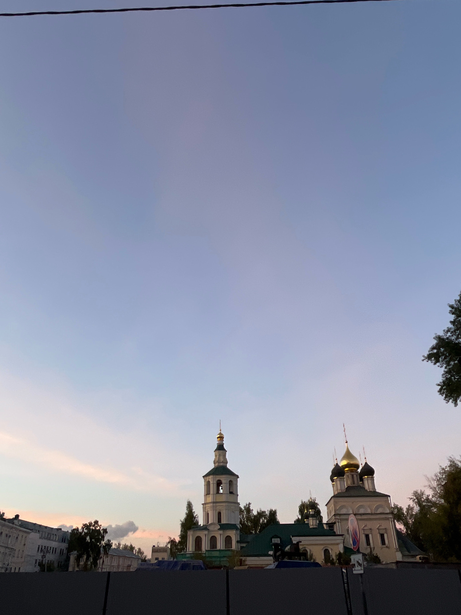 The image captures a view of the Church of the Resurrection in Kadashi, located in Moscow, Russia, at dusk. The sky is a gradient of pale blue to soft pink, indicating the setting sun. A wire runs horizontally across the upper portion of the frame. In the foreground, a dark fence obscures the lower part of the scene. Behind the fence, the Church of the Resurrection is visible, with its white and green bell tower topped by a golden dome. Adjacent to it is another section of the church with multiple golden domes. Trees are scattered around the church structures. To the left, a row of apartment buildings with light-colored facades is visible. There are no people or animals visible in the image.
