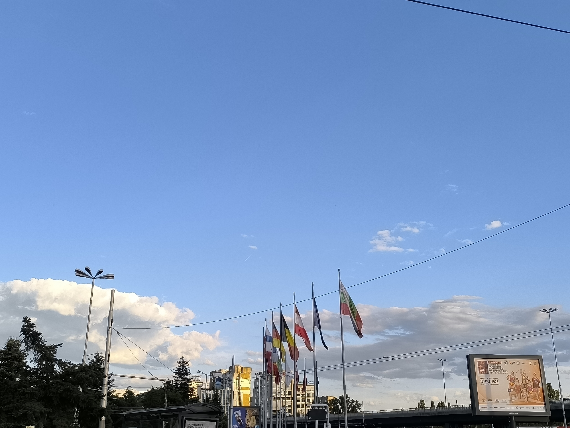 The image depicts a street scene in Sofia, Bulgaria, on a clear day with scattered clouds. In the foreground, several flags are displayed on flagpoles, including the Bulgarian flag. Trees and streetlights are also visible. In the background, buildings and an overpass can be seen. A billboard is prominently featured on the right side of the image, displaying information about an event. The overall impression is of an urban environment with a sense of civic pride indicated by the flags.