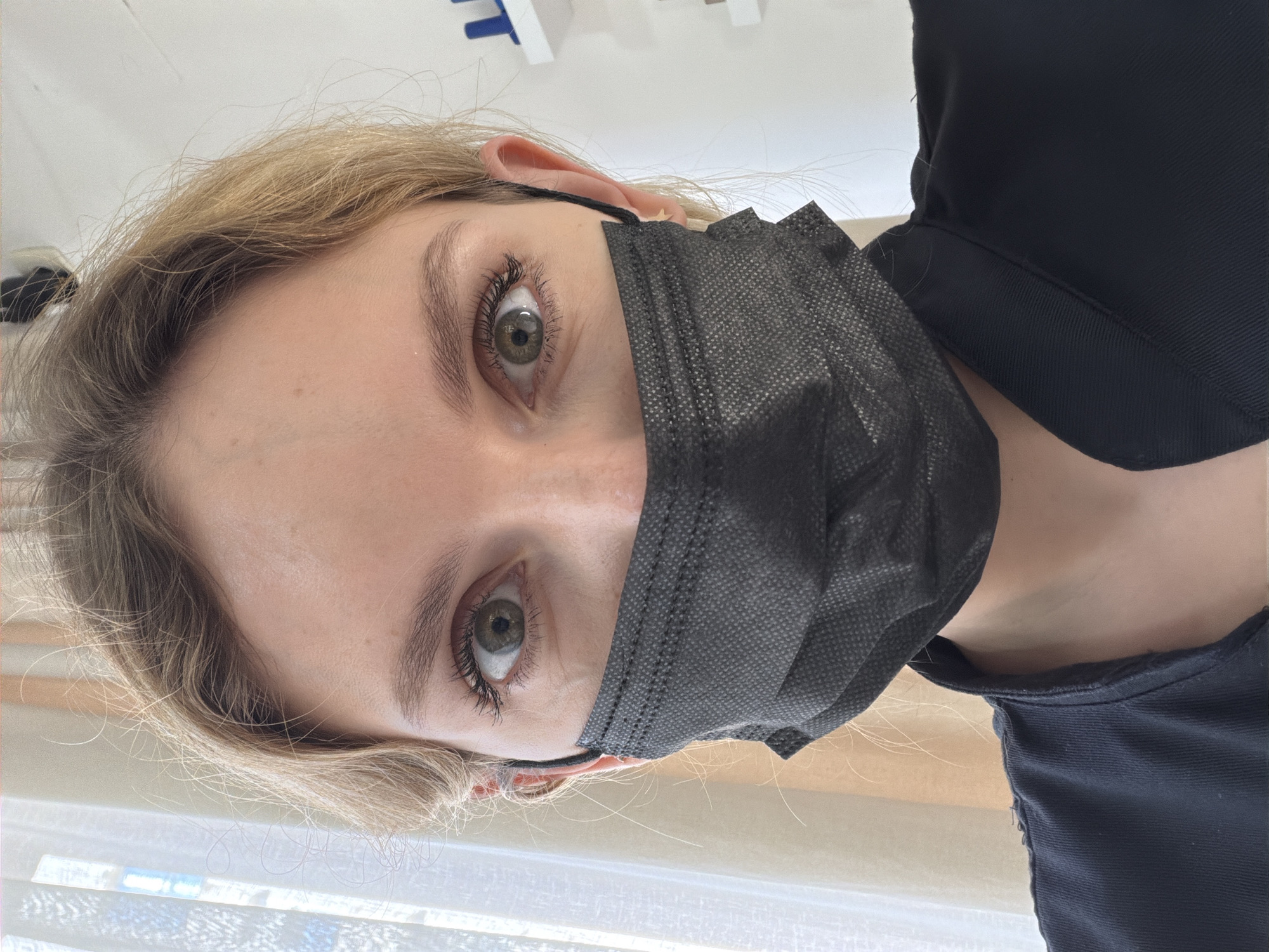 A vertically oriented close-up image depicts a person wearing a black face mask. The person has blonde hair, light skin, and green eyes. Part of their right ear is visible, and they are wearing a dark-colored garment. The background appears to be a neutral-colored interior space, possibly near a window with blinds. There is no evidence to suggest this image was taken in Budva, Montenegro.