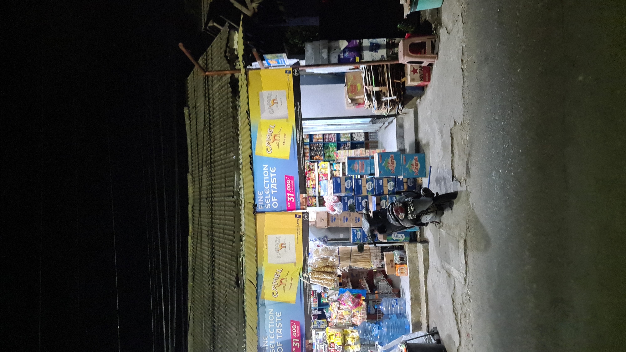 The image shows the storefront of a small convenience store or minimarket at night in Denpasar, Indonesia. The store is brightly lit with banners advertising "Camel Fine Selection of Taste" cigarettes for Rp 31,000. Various goods are displayed, including bottled water (Aqua, Le Minerale), snacks, and what appear to be packaged food items. A motorcycle is parked in front of the store. The surrounding area appears to be a street with a worn concrete surface and a tiled roof overhead. Electrical wires are visible above the store. The overall scene suggests a typical street-level retail establishment operating after dark.