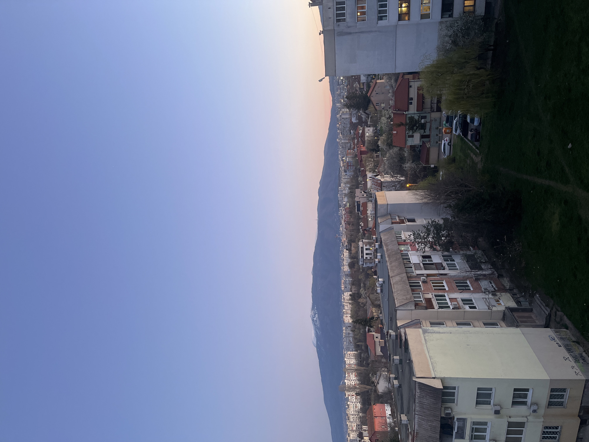 Here's a detailed description of the image:
**Setting:** An elevated view of Sofia, Bulgaria, taken at dusk. The sky transitions from deep blue to a faint orange glow near the horizon, suggesting either sunrise or sunset.
**Environment:** The shot overlooks a densely populated urban area. Buildings of varying heights and architectural styles dominate the landscape, mostly residential apartment buildings. Between the buildings there are trees, bare trees are more visible with the trees having green leaves not so visible. The area is bordered by the Vitosha mountain, visible in the background with snow visible on some peaks.
**Buildings:** A mix of aging, Soviet-era apartment blocks and newer constructions. Some buildings feature red-tiled roofs.
**Details:** Cars are parked in designated areas. In the sky there are no clouds and no objects.