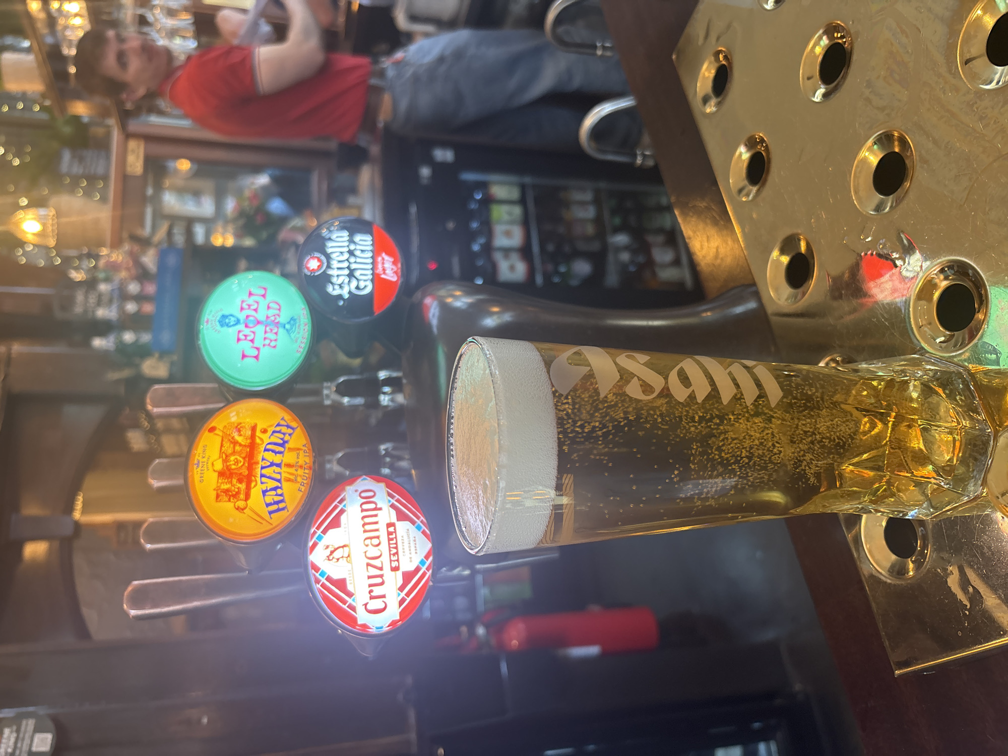 The image shows a close-up of a bar with a row of beer taps and a glass of beer. The taps are labeled with different beer brands, including "LEBEL HEAD", "Estrella Galicia", "HAZY DAY", and "Cruzcampo". A glass of "Asam" beer sits in front of the taps. A man stands at the bar, wearing a red t-shirt. The background is a blur of lights and other patrons. The scene is possibly set in a pub or bar in London, United Kingdom.