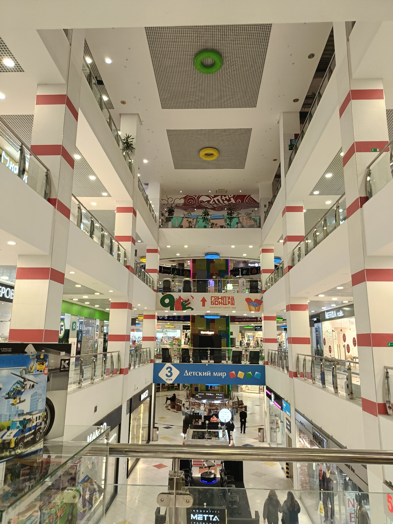Here is a description of the image:
The image shows the interior of a multi-level shopping mall, possibly located in Saint Petersburg, Russia, based on the Cyrillic script seen in some signage. The architecture is modern, with white pillars accented by horizontal red stripes. There are open spaces between floors, creating a visual connection between levels. Clear glass railings offer views down to lower levels, where various shops are visible.
Signage for "Детский мир" (Detsky Mir), which translates to "Children's World," can be seen, along with other signs with Russian text. There are also recognizable brand names such as "MANGO." People can be seen walking on the ground level. The ceiling has a grid-like design with round, decorative elements in green and yellow. Overall, the scene depicts a brightly lit and bustling shopping environment.