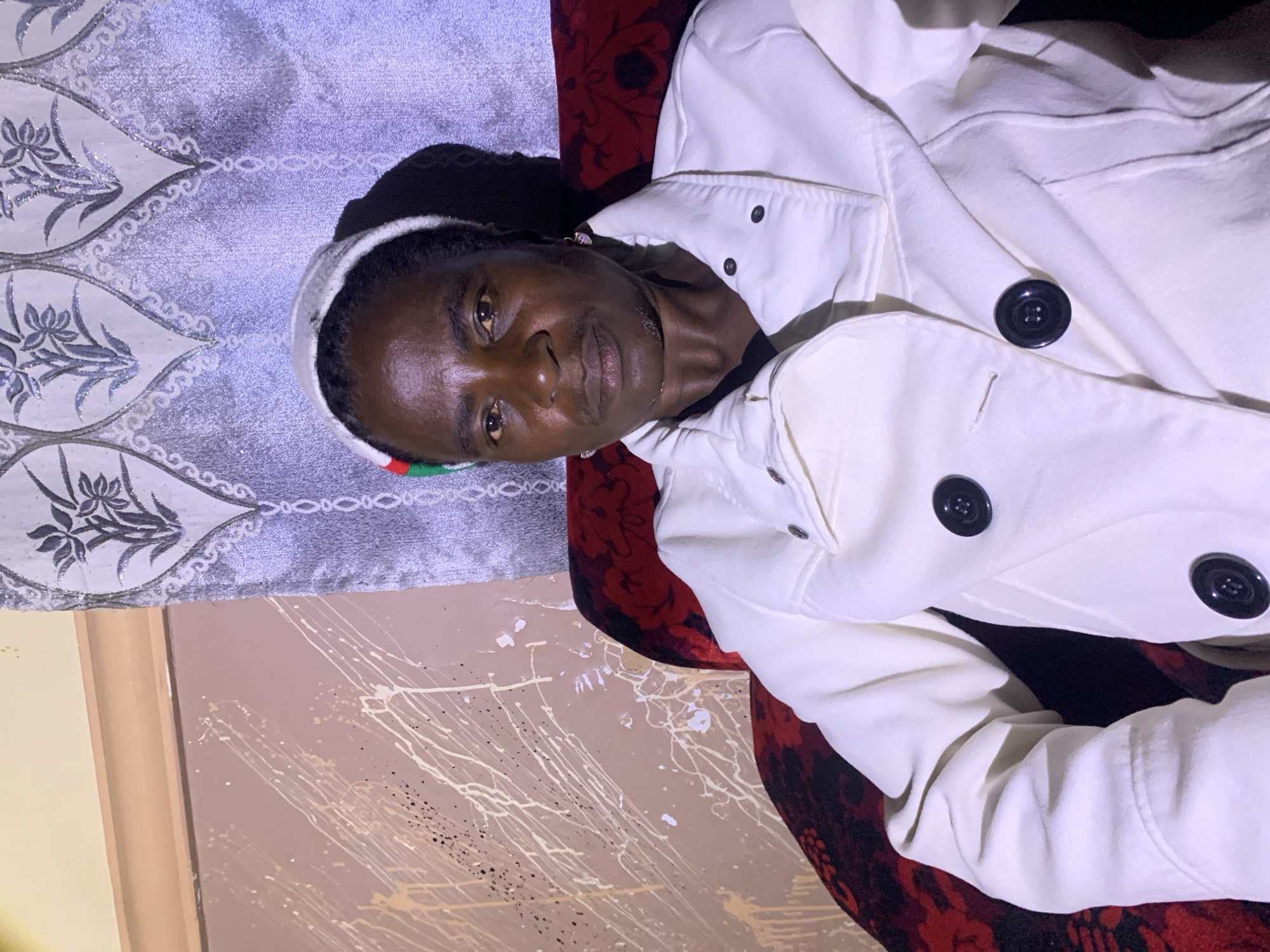 A middle-aged Black woman with dark skin and short, dark hair is sitting in a red armchair. She is wearing a white coat with black buttons and a white cap with a red, green, and black stripe on the front. She is looking directly at the camera with a neutral expression. Behind her, there is a wall with a light brown, textured paint finish with white and cream splatters. To the right of the wall is a gray curtain with a floral pattern. The lighting suggests an indoor setting, possibly a home, in Barakin Sabawa, Nigeria.