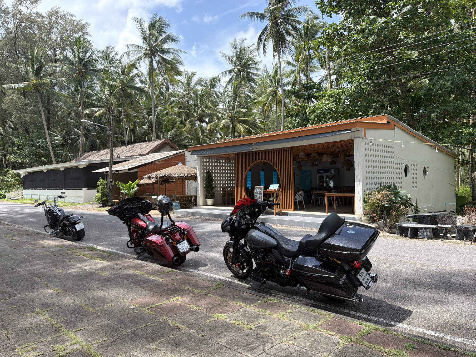 Three motorcycles are parked on the side of a road in front of a building with an open-air seating area, located in Bang Muang, Thailand. The motorcycles are positioned on the pavement and the road. The first motorcycle, a black cruiser, is parked on the road to the left, facing away from the camera. The second motorcycle, a dark red cruiser, is parked on the road to the left, angled towards the camera, with a black helmet resting on its rear. The third motorcycle, a dark grey cruiser, is parked on the road to the right, facing towards the camera, also with a black helmet on its rear. The building appears to be a cafe or restaurant, with tables and chairs visible inside and outside. Palm trees and other tropical foliage surround the scene, suggesting a warm, sunny day with a clear blue sky. The road surface is asphalt, and the pavement is made of large, rectangular tiles.