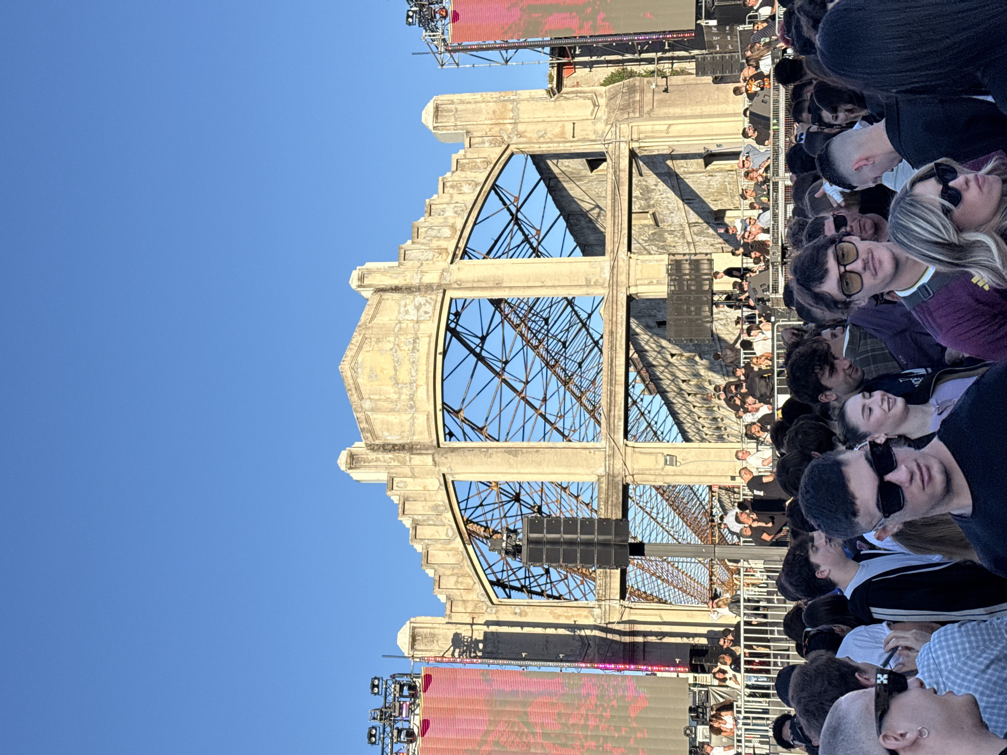 The image shows a large crowd of people gathered in front of a dilapidated structure, possibly a remnant of an old industrial building. The structure is made of concrete and steel, with large arched openings and a visible internal framework. Speakers and a large LED screen are positioned in front of the building, suggesting a concert or event is taking place. The sky is clear blue, and the photo appears to be taken during the day. Based on the architectural style and the presence of the abandoned factory structure, it is likely the location is in Milan, Italy, possibly within or near the Parco Nord Milano, a large urban park where abandoned factories are sometimes used for events.