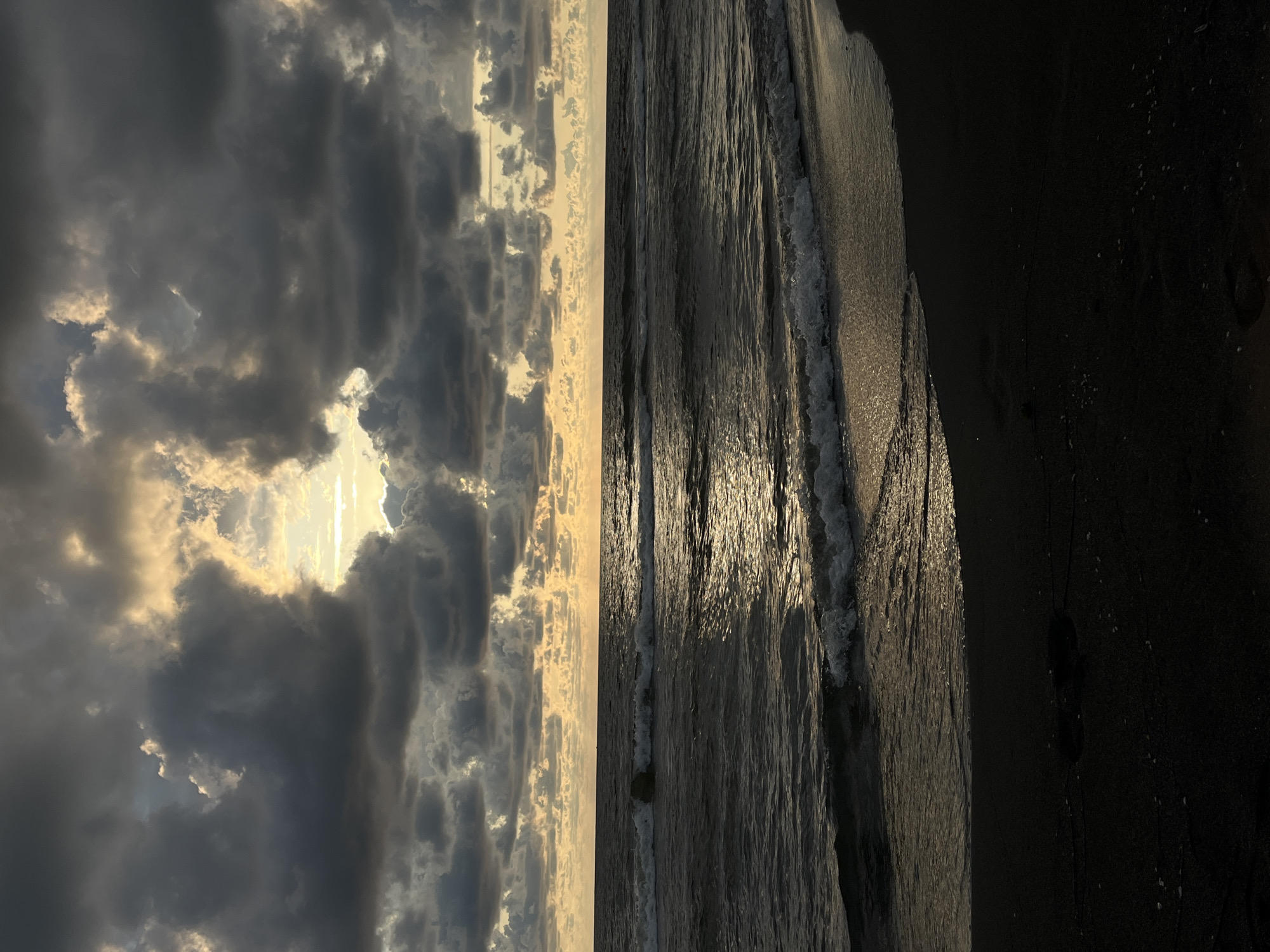 This is a vertical, high-angle shot of a beach at sunset. The image is divided into two distinct halves. The left side features a dramatic sky filled with dark, heavy clouds illuminated by bright streaks of sunlight peeking through. The right side shows the dark sand of the beach with gentle waves lapping at the shore. The water reflects the golden light of the setting sun, creating a shimmering effect on the surface. Footprints are visible in the wet sand near the water's edge. The overall mood is serene and contemplative, with the dramatic sky contrasting with the calm sea. The location is given as Sufs Community, Georgia. The time of day is sunset, and the weather appears to be clearing after a potential storm, with dramatic cloud formations and sunlight. There is no visible text in the image.