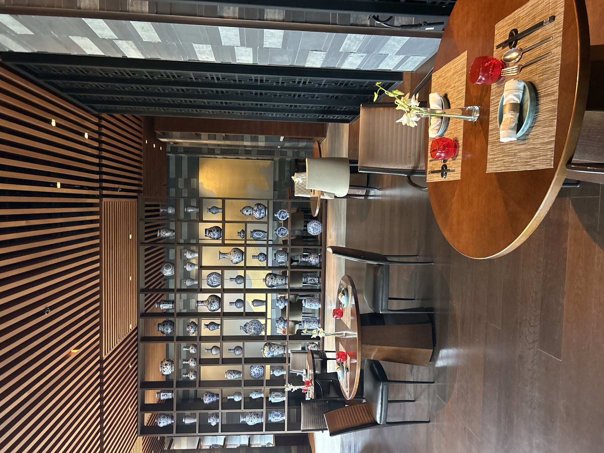 This is a photograph of a dining area with traditional Chinese-inspired decor. The room features a tiered shelving unit filled with blue and white porcelain vases and pottery. The ceiling is adorned with wooden slats, and the walls are a mix of stone and decorative screens. Several dining tables are set with placemats, cutlery, red glasses, and small floral arrangements. The flooring is dark wood. The image appears to be taken indoors, likely in a restaurant, with no people visible. This dining area is located in a restaurant in Pune, India.