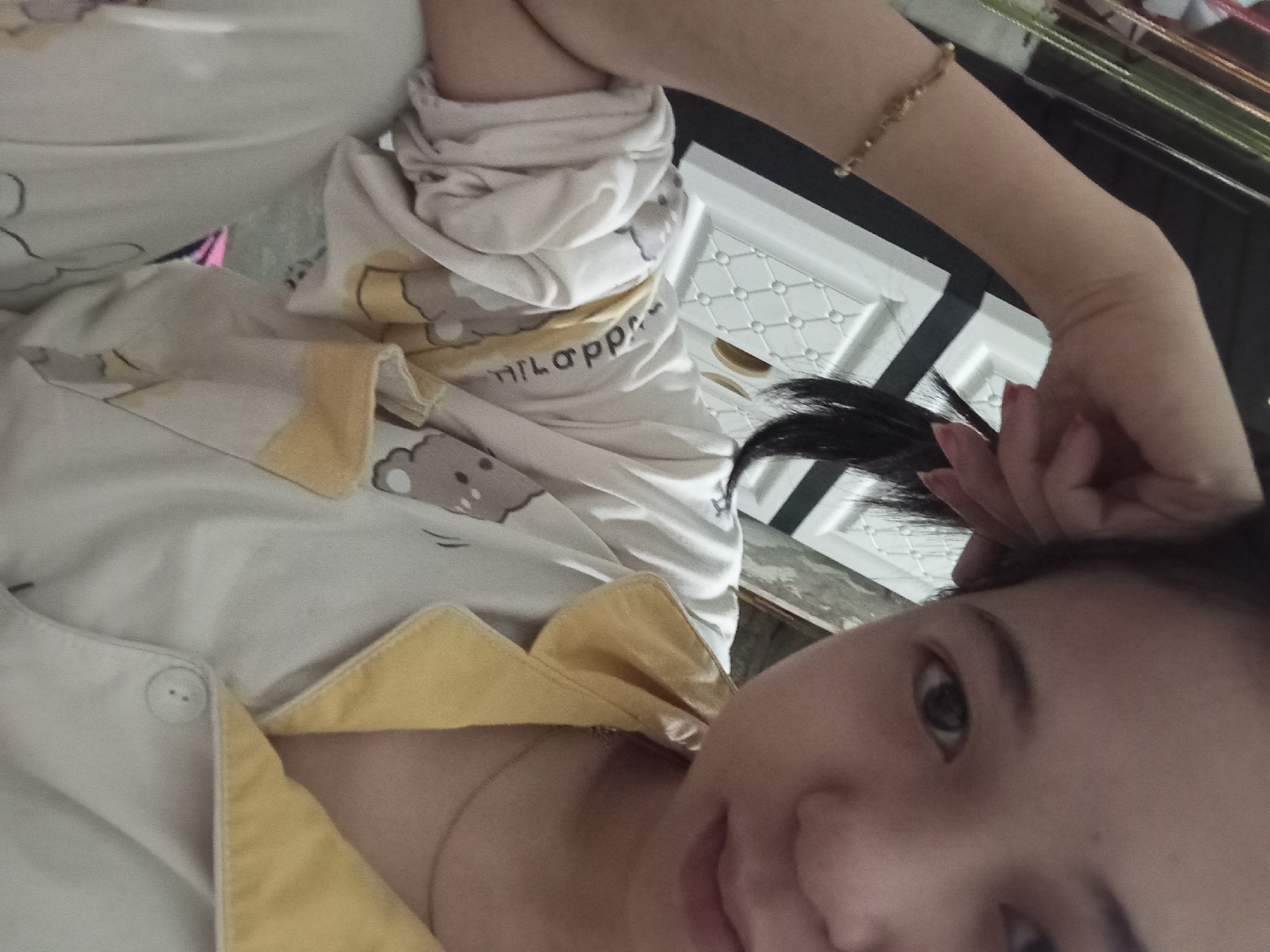 A young woman is taking a selfie indoors. She is wearing light-colored pajamas with a yellow collar and a pattern of cartoon bears. Her left arm is raised, and she is touching her hair with her hand. A beaded bracelet is visible on her left wrist. The background includes a white cabinet with a diamond pattern and some shelving units. The scene appears to be in a bedroom or personal living space, likely within a residence in Rejosari, Indonesia.