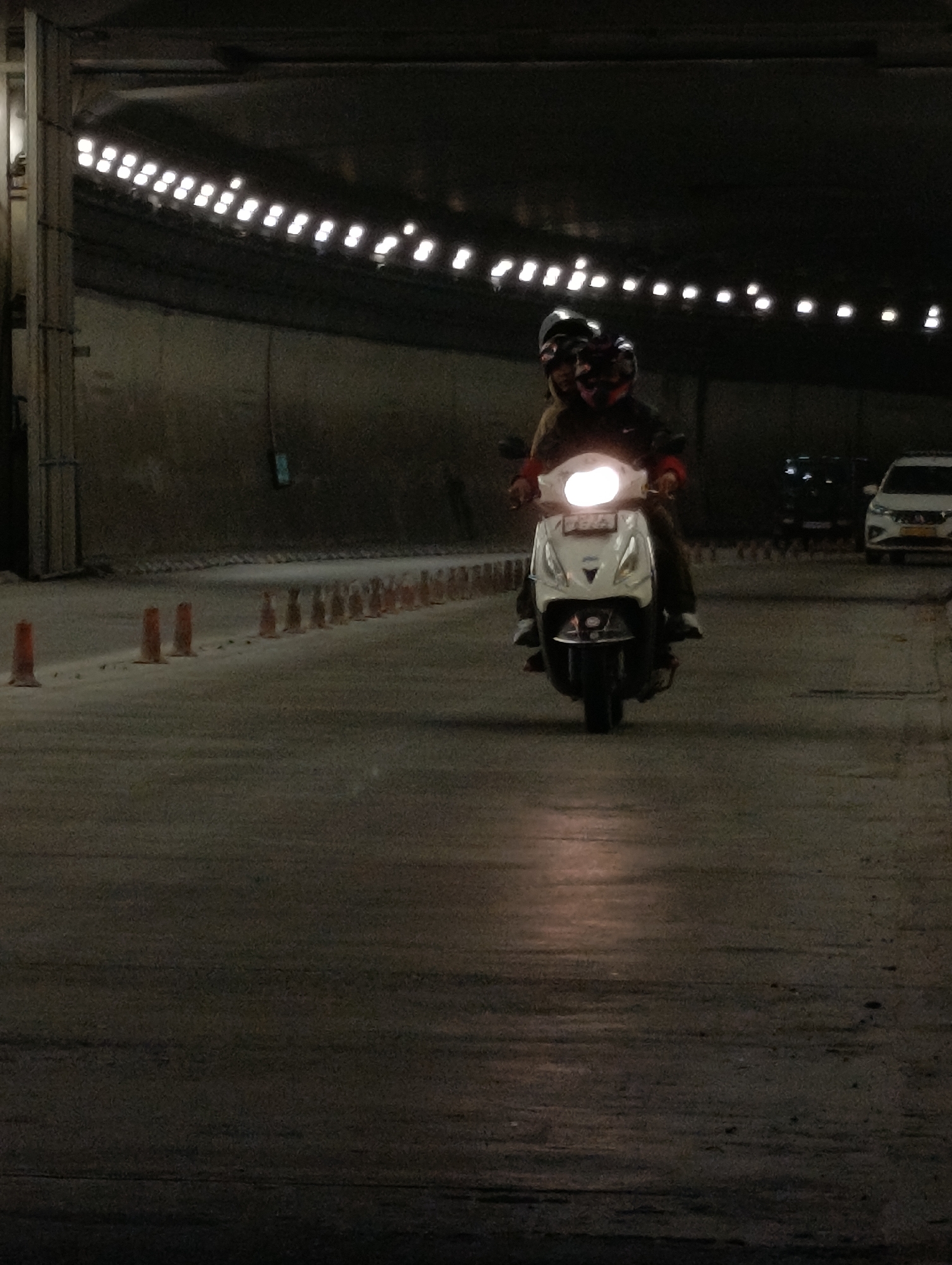 Two individuals are riding a white scooter inside a dimly lit tunnel. The rider in front is wearing a helmet and a light-colored jacket, while the passenger behind is wearing a helmet and a dark-colored jacket. The scooter's headlight is brightly illuminated. In the background, a white car is visible, and a line of orange traffic cones is present on the right side of the road. The tunnel walls are concrete, and a row of lights runs along the ceiling. The road surface appears wet and reflective, suggesting recent rain or a damp environment. The scene takes place in the city of Unknown, India.