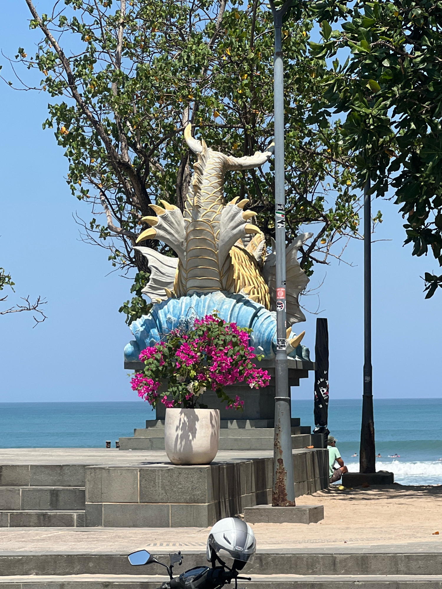 The image depicts a scene in Kuta, Indonesia, specifically on a sunlit beach promenade. In the foreground, the front of a white and silver motorcycle helmet is visible on a set of concrete stairs. Behind the helmet, the promenade itself is constructed of stone and concrete steps leading up to a raised area.
Dominating the middle ground is a large, ornate statue of a mythical creature, possibly a dragon or serpent, rendered in shades of white, gold, and blue. The statue is seated on a blue, wave-like base and is adorned with bright pink bougainvillea flowers in a large, light-colored pot placed in front of it. Several trees with green foliage provide a natural backdrop, their branches reaching towards a clear blue sky. A tall, grey lamppost stands to the right of the statue, with stickers adhered to its surface. Another, thinner lamppost is visible further to the right.
In the background, the turquoise waters of the ocean stretch to the horizon, with gentle waves breaking on the sandy beach. A few people can be seen on the beach; one individual wearing a light green shirt and shorts sits on the sand, facing the ocean, while others are in the water, seemingly engaged in surfing. The overall atmosphere is one of a vibrant, tropical coastal setting.