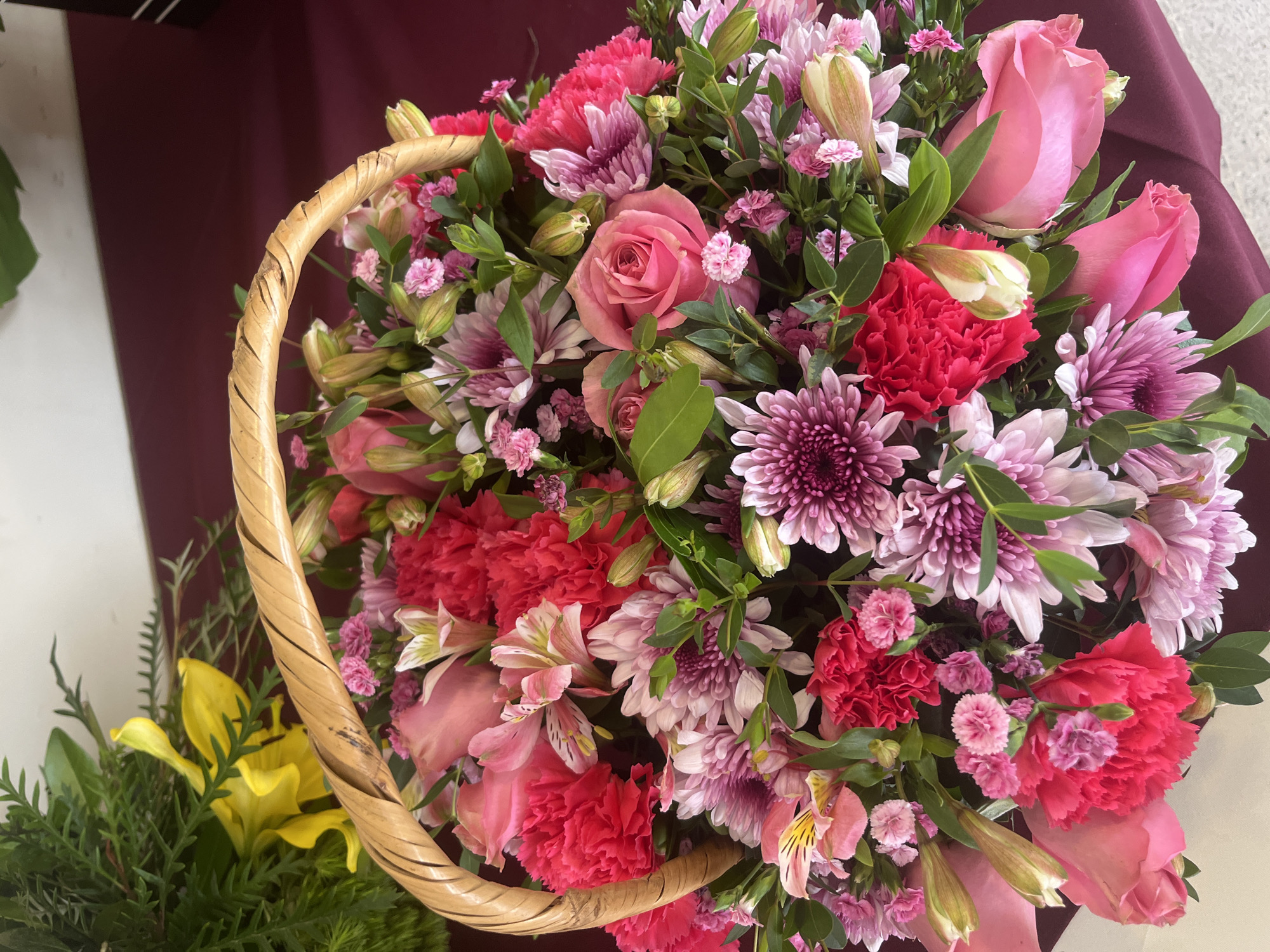 A high-angle shot depicts a wicker basket overflowing with a bouquet of flowers in a variety of pink, red, and purple hues. The floral arrangement includes roses, carnations, chrysanthemums, and lilies, interspersed with green foliage. A patch of yellow flowers is visible to the left of the basket. The bouquet rests on a maroon-colored surface. The location is possibly in Puebla, Mexico, but there is no specific evidence within the image to confirm this.