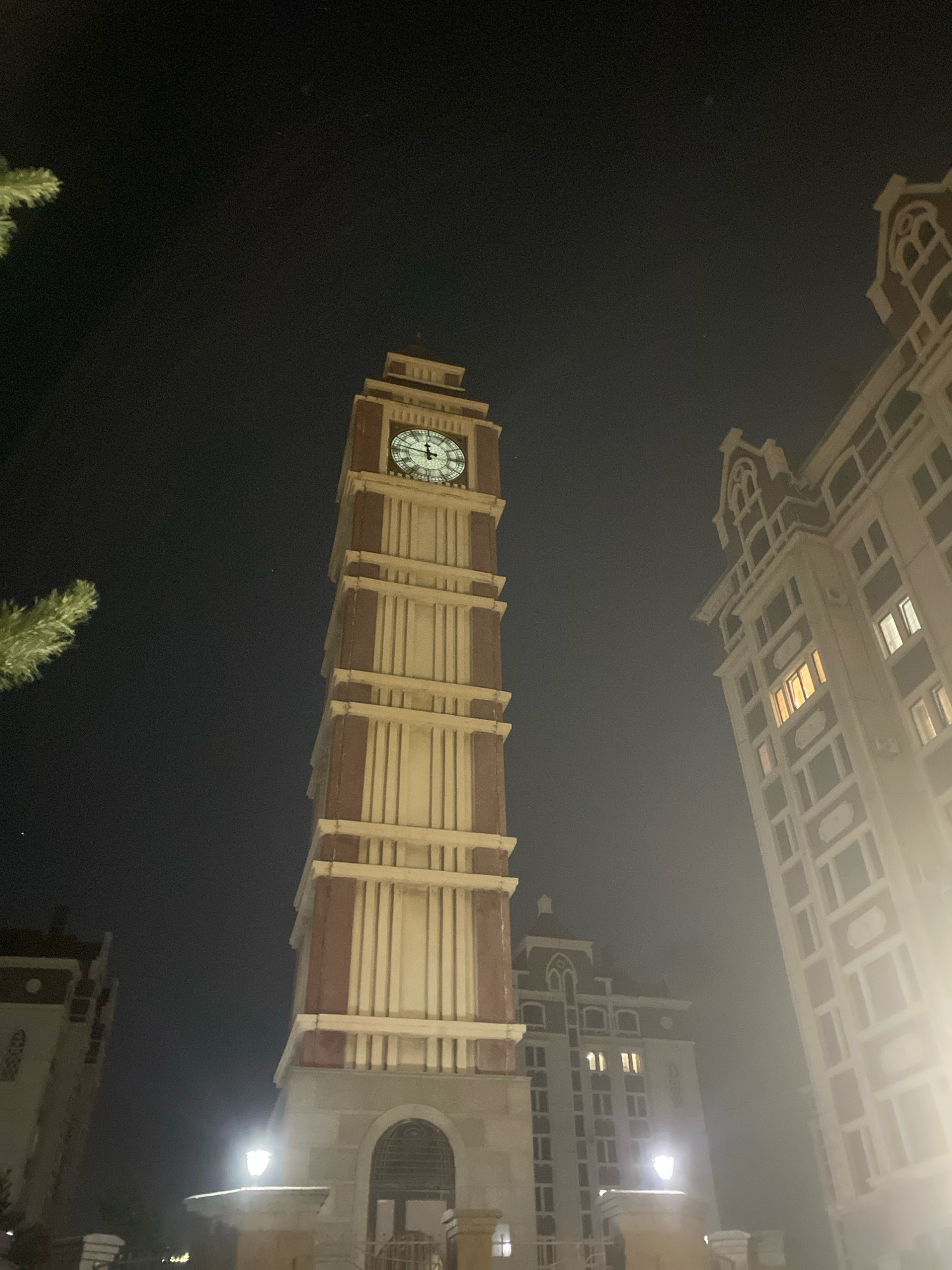 A nighttime, low-angle, wide shot captures the exterior of a clock tower and surrounding buildings in Astana, Kazakhstan. The central focus is a tall, multi-story clock tower with a large, illuminated clock face showing the time as approximately 3:05. The tower is constructed with light-colored vertical elements framed by reddish-brown bands. To the right of the clock tower, a tall, multi-story building with numerous windows is visible, some of which are lit. Another building, with a distinctive architectural style featuring arched windows, is partially seen behind and to the left of the clock tower. A branch of a green, feathery tree is in the upper left corner of the frame. The sky is dark and hazy, indicating nighttime conditions. Streetlights are visible at the base of the clock tower, casting light onto the surrounding area. The overall impression is of an urban nighttime scene.