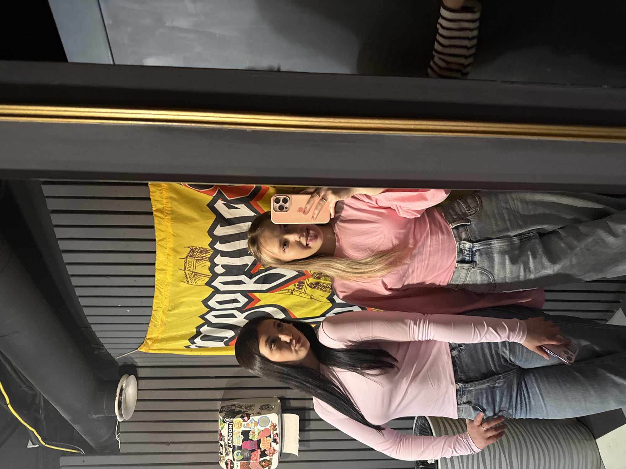 The scene depicts two young adult females posing for a mirror selfie in an interior setting located in Chaikovsky, Russia, likely a restroom or changing area of a commercial establishment.
On the left, a woman with long dark hair, wearing a long-sleeved light pink top and light-wash blue jeans, stands facing slightly right with her head tilted and a pouty expression. Her right hand is on her hip, and her left hand holds a pink-cased smartphone near her thigh. To her right, a second woman with long blonde hair, also wearing a long-sleeved light pink top and light-wash blue jeans, is reflected in a large, dark-framed mirror with a gold-colored inner trim. She is actively taking the selfie, holding a pink iPhone 13 Pro Max to her face with her right hand and sticking out her tongue. A partial reflection of another individual's arm, wearing a black and white striped long-sleeved shirt, is visible in the far right of the mirror.
The background consists of dark gray vertical wall paneling. Above the women, exposed black pipes run along the black ceiling, accompanied by yellow electrical conduit and a white, round, industrial-style light fixture. A white, wall-mounted toilet paper dispenser covered in various stickers, including one that reads "LAGOON," is visible on the left wall, with a roll of toilet paper hanging from it. A large yellow flag with a black gothic-style inscription (partially visible as "SPORTL...") and an illustrative drawing of a building or ship structure is hung on the wall behind the women. A vertically ribbed silver or gray trash receptacle is partially visible in the bottom-left foreground.