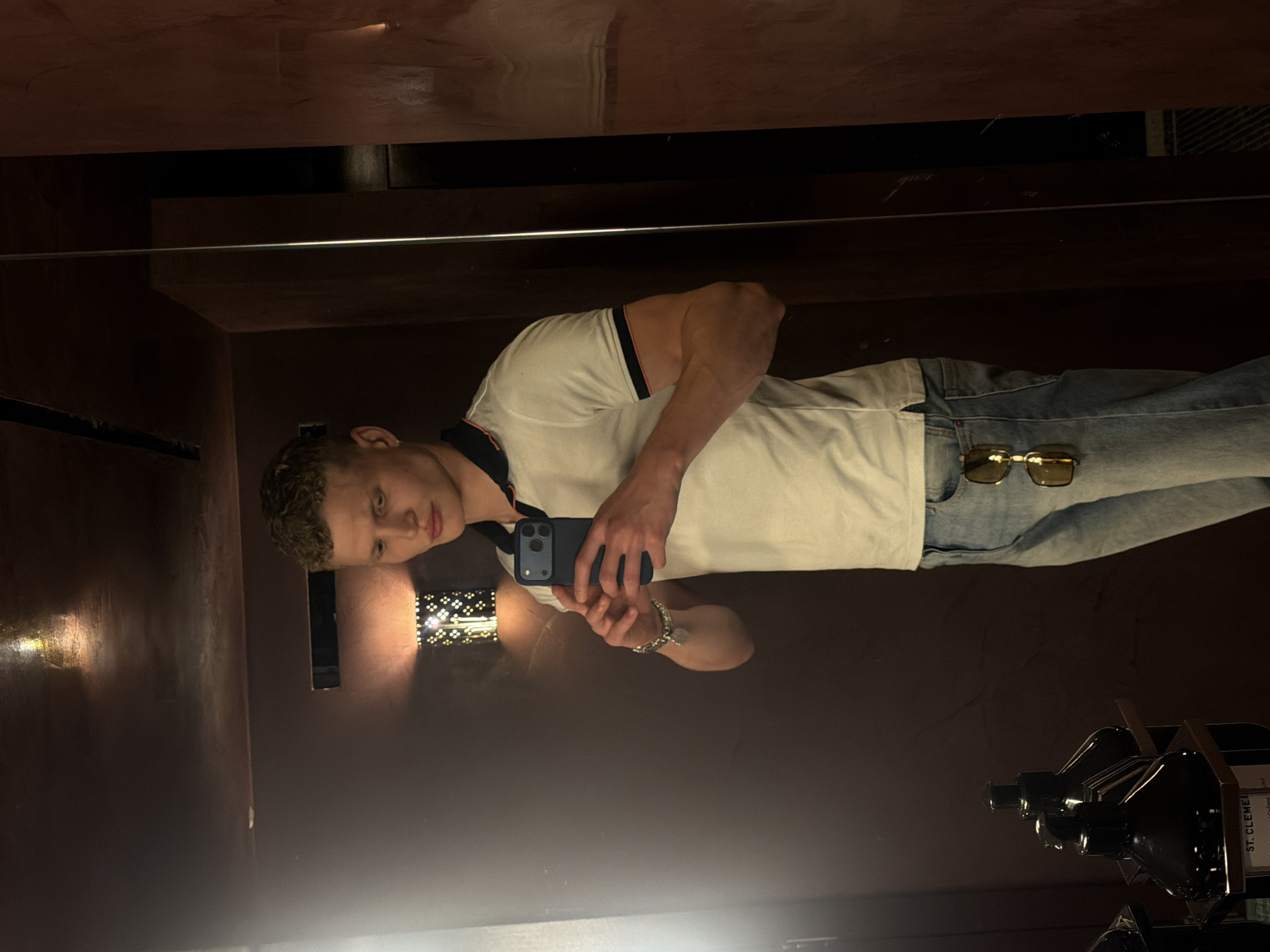 A young man with curly blonde hair stands in front of a dark, textured wall and a mirror, taking a selfie with his phone. He is wearing a white polo shirt with blue and red trim on the collar and sleeves, and light-wash denim jeans. A pair of sunglasses is hanging from the front of his jeans. The environment appears to be a restroom or dressing room, with a dimly lit wall fixture emitting a patterned light, and what seem to be bottles of toiletries on a shelf in the lower left corner of the frame. The specific location within London, United Kingdom, cannot be determined from the visual information provided.
