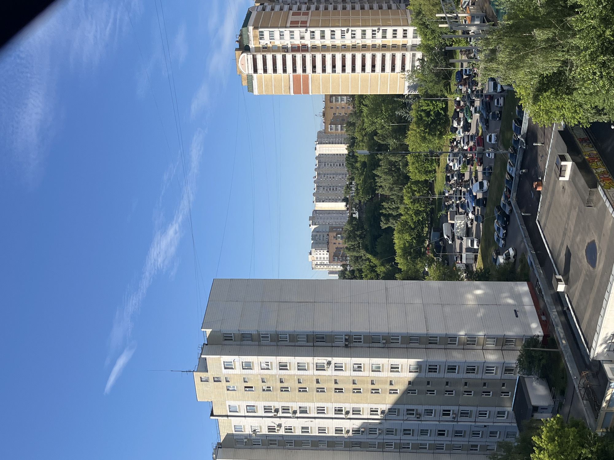The image captures a panoramic view of a residential area in Moscow, Russia, on a clear day. In the foreground, two multi-story apartment buildings dominate the scene. The building on the left is a tall, rectangular structure with a light-colored facade and numerous windows. The building on the right is also a high-rise, distinguished by its yellow and white exterior with visible balconies. Between these buildings and in the background, a dense canopy of green trees forms a lush, natural buffer. Beyond the trees, several other apartment complexes with similar architectural styles are visible, characteristic of Soviet-era urban planning. A parking lot filled with numerous cars is situated in the middle ground, indicating the presence of residents. Power lines crisscross the sky, a common sight in urban environments. The sky is a vibrant blue with a few wispy clouds, suggesting fair weather conditions. The overall impression is one of a typical urban landscape in a large Russian city, characterized by a mix of high-density housing and green spaces.