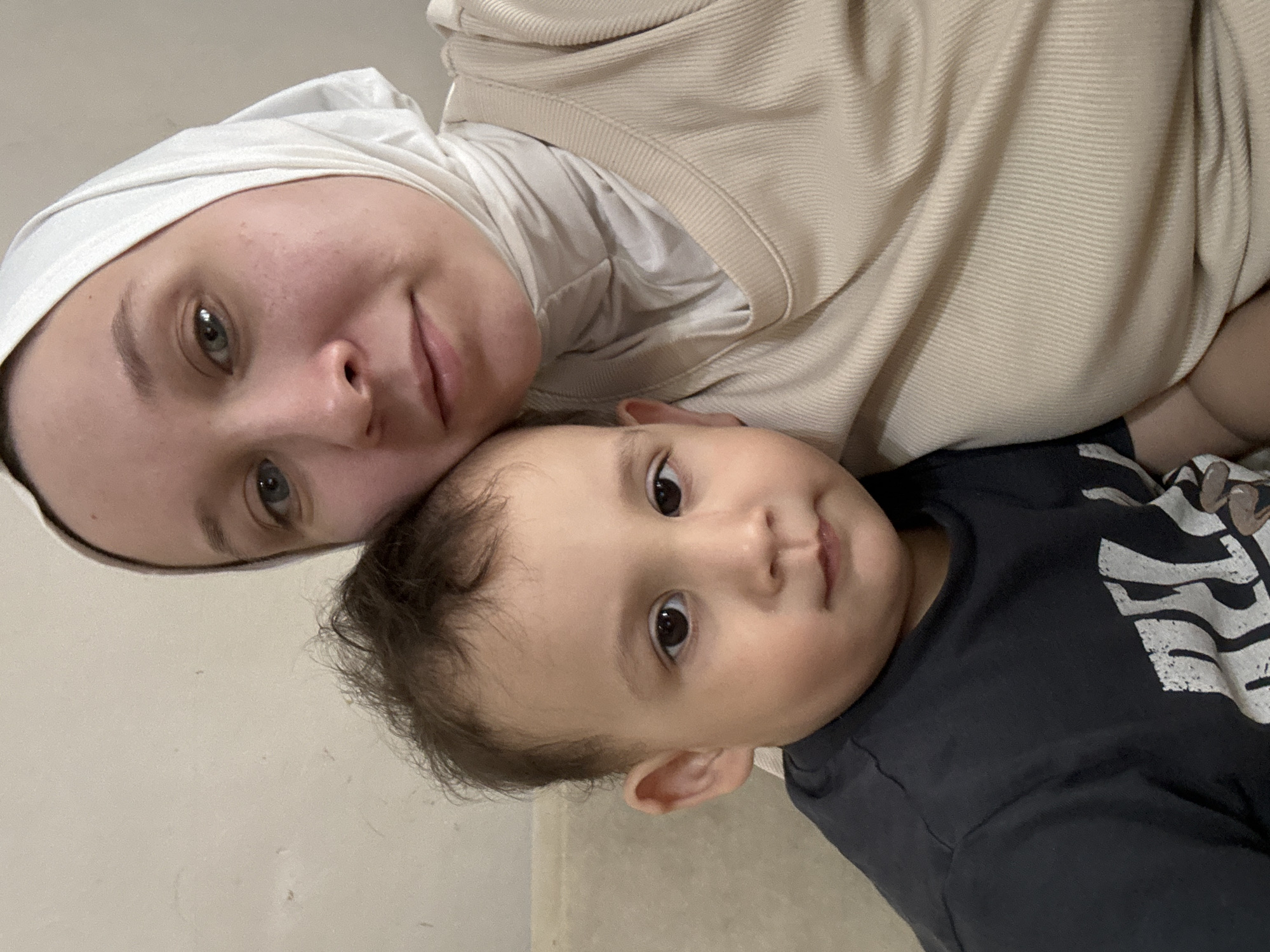 A woman wearing a white headscarf (hijab) and a beige sweater is shown with a young child wearing a black t-shirt with a white graphic. They are lying down, with the woman's head near the top-left of the frame and the child's head near the center. The background appears to be a neutral-toned wall or floor. The location is specified to be in Chaikovsky, Russia.