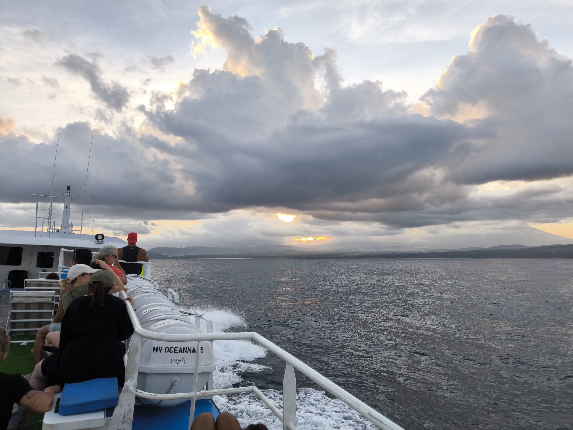 The photo depicts a scene from a boat, presumably in Unknown, Indonesia, based on the presence of Mount Agung in the background. The boat is identified as "MV Ocean 9" on a white cylindrical object. Several people are on the boat, with their backs to the camera. A man in a red hat and black tank top stands at the rail. In the distance, land is visible beneath a dramatic sky filled with large, dark clouds, through which the sun is setting or rising. The water is choppy, creating white foam as the boat moves. The overall light is subdued due to the heavy cloud cover.