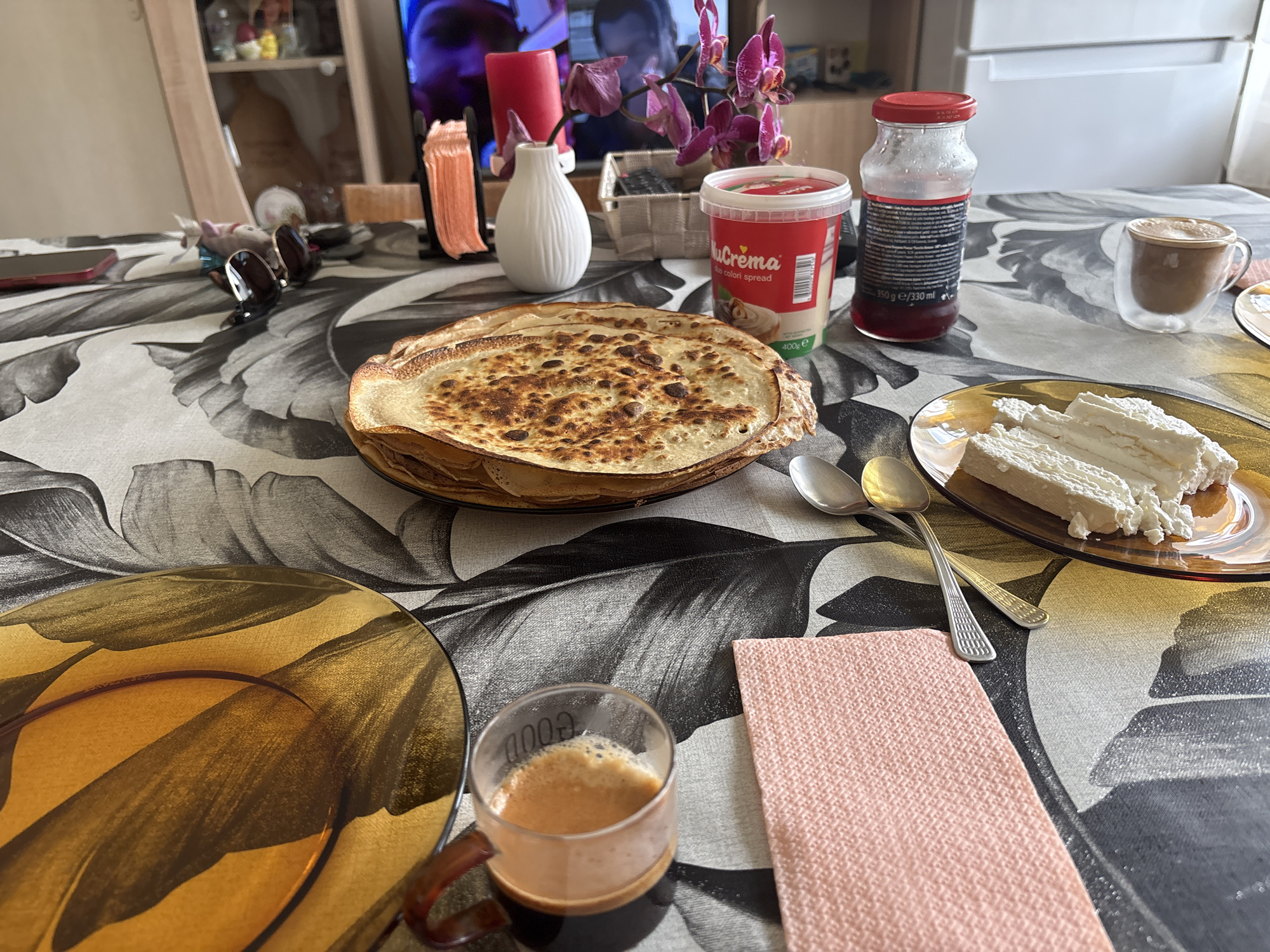 The image depicts a close-up view of a table set for breakfast, likely in an indoor setting. A stack of golden-brown crepes is prominently displayed on a dark plate in the center of the frame. To the right of the crepes, a small glass cup contains a frothy, brown beverage, possibly coffee or espresso, with the word "GOOD" visible on its side. A pink napkin lies to the right of the coffee cup. Further to the right, a thin slice of white cake or pastry, possibly topped with cream cheese or ricotta, rests on an amber glass plate, accompanied by two silver spoons. In the background, to the left, a red cylindrical object and a white vase with purple orchids add decorative elements. A red plastic container labeled "NuCrema" is visible near the crepes. To the right of the NuCrema container, a glass bottle with a red cap contains a dark red liquid. The table is covered with a tablecloth featuring a large, abstract floral or leaf pattern in black and white. The scene does not contain any specific visual cues that would place it in Gabrovo, Bulgaria.