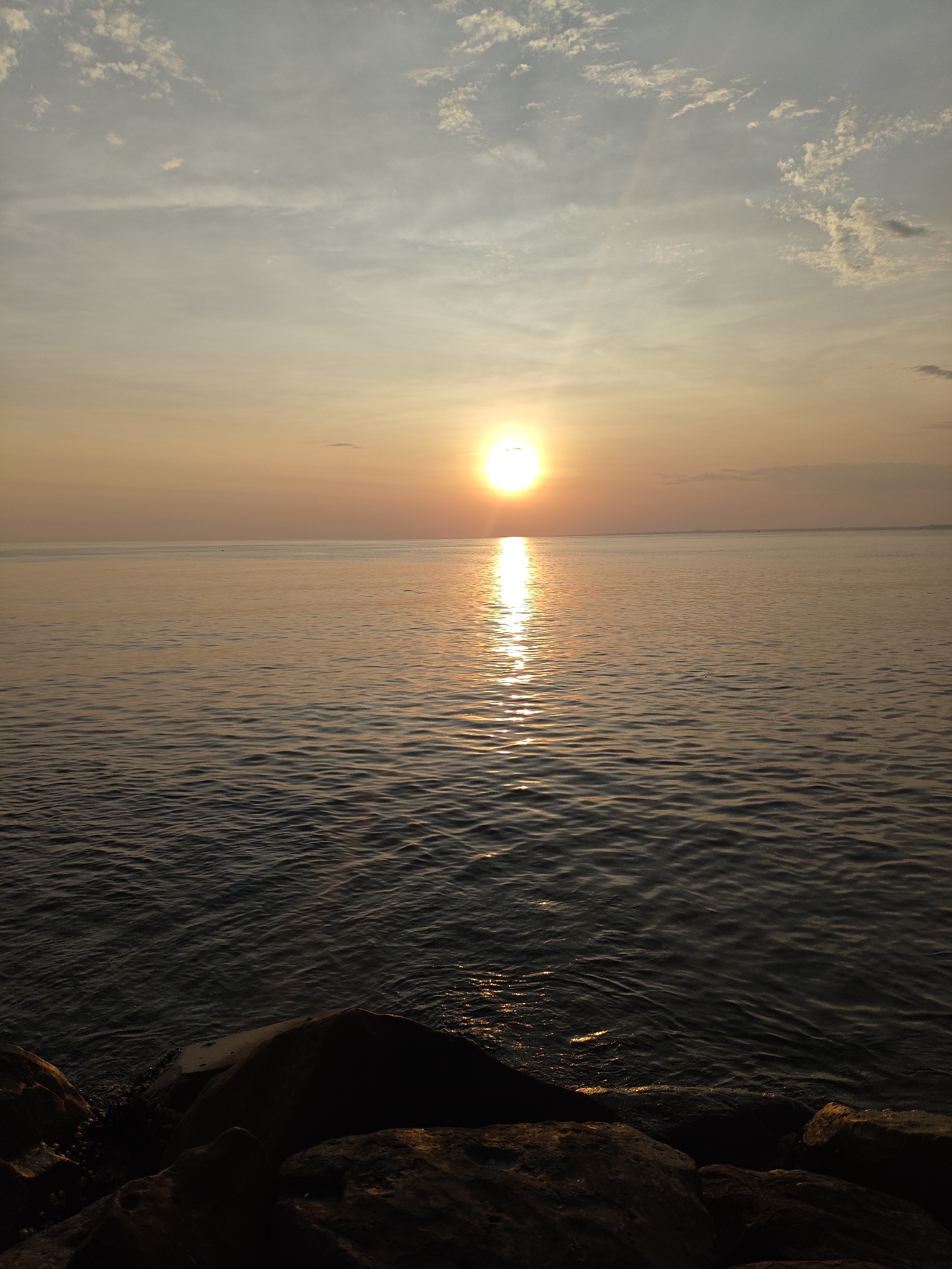 The image displays a coastal landscape in Lhokseumawe, Indonesia, captured during sunset. The sun, a bright yellow-orange sphere, is positioned low above the horizon, slightly to the left of the frame's vertical centerline. Its intense light generates a shimmering, undulating vertical reflection across the water's surface. The sky transitions from a pale blue-grey at the top to a warm amber-orange near the horizon, containing scattered, white, small cloud formations. The expansive body of water, likely the sea, exhibits gentle ripples and reflects the sun's light. The immediate foreground is occupied by several large, dark, irregularly shaped rocks, partially submerged, forming a rocky shoreline. No individuals, vessels, or other specific objects or actions are discernible within the frame.