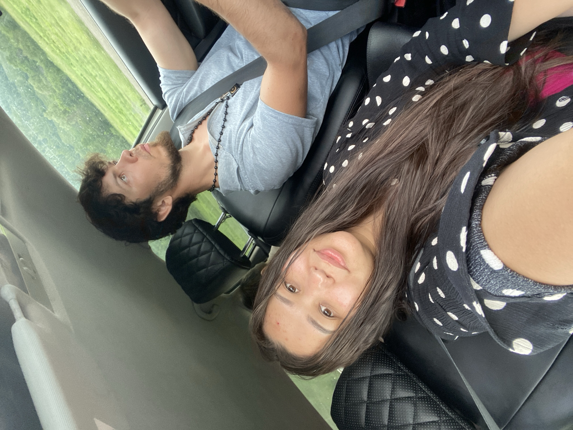 The media file shows two individuals, a man and a woman, seated in the front passenger seats of a car. The man, on the left, is looking upwards and to the left, with a beard and dark curly hair. He is wearing a grey t-shirt and a black beaded necklace. A seatbelt is visible across his chest. The woman, on the right, is facing the camera, with long brown hair and brown eyes. She is wearing a black and white polka dot top. A seatbelt is also visible across her body. The interior of the car appears to be black leather. Outside the car window, a green, slightly blurry landscape is visible, suggesting a rural or natural setting. The lighting suggests it is daytime. No text is visible in the image. The orientation of the image is rotated.