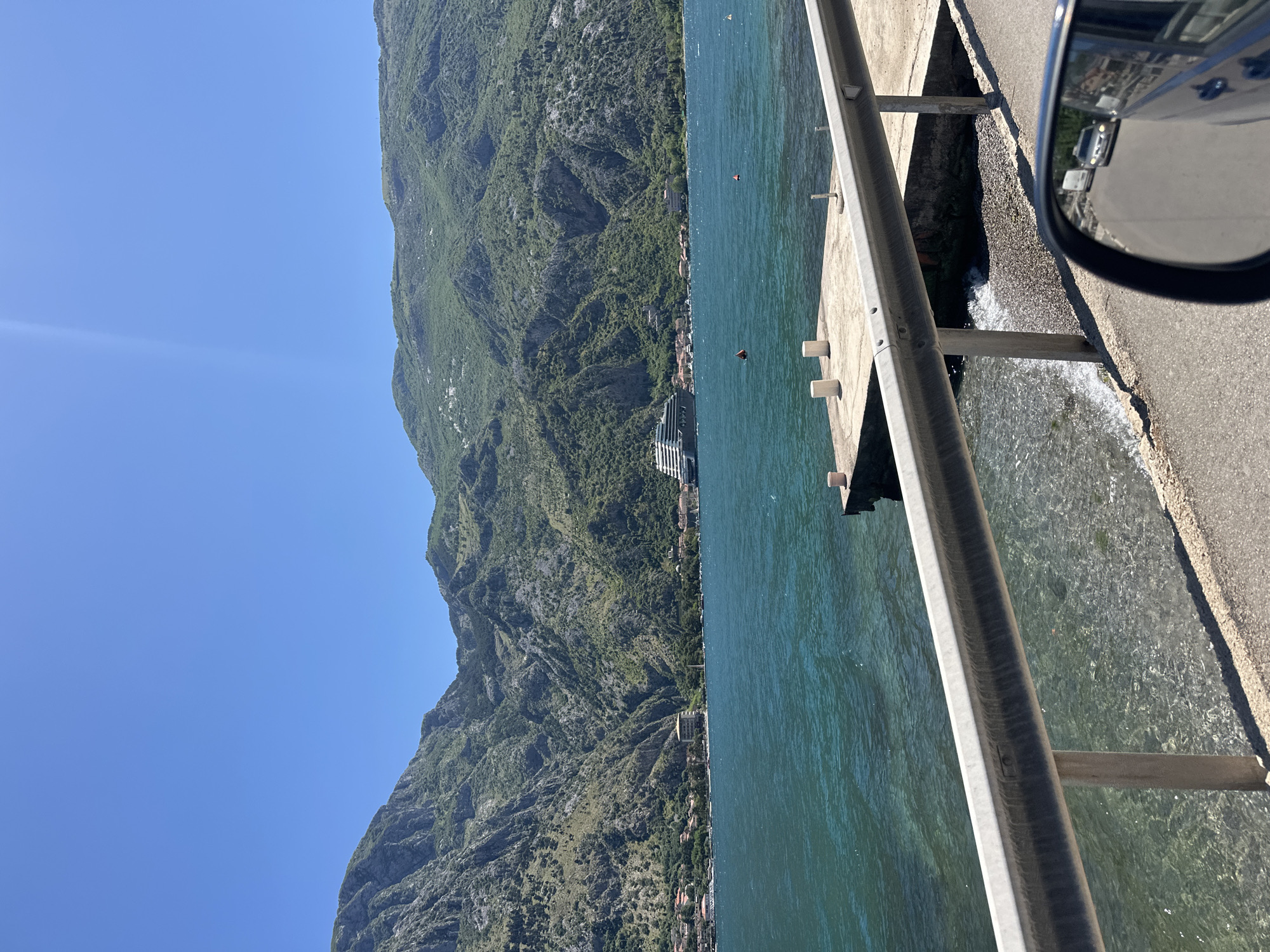 View of the Bay of Kotor from a road in Prčanj, Montenegro. A mountain covered in lush green vegetation rises from the shore on the left. The blue water of the bay occupies the center of the frame. A grey metal guardrail runs parallel to the road, which is made of concrete or asphalt. Part of a car, specifically a side mirror, is visible on the right edge of the frame, reflecting the environment and indicating that the photo was taken from inside a vehicle.