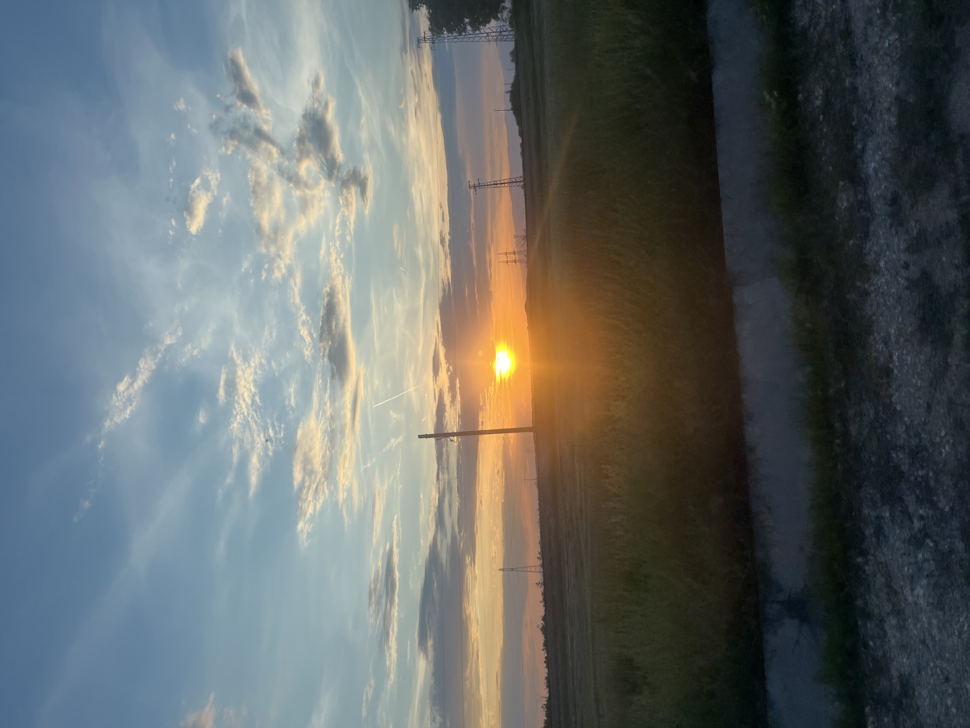 The image depicts a landscape, likely in Dolna Mahala, Bulgaria, at sunset. The sun is positioned near the horizon, casting a bright orange glow across the scene. Power lines and utility poles stretch across the landscape. To the right of the frame is a dark path, and beyond that appears to be a field. The sky is partially cloudy with hues of blue and orange from the setting sun.