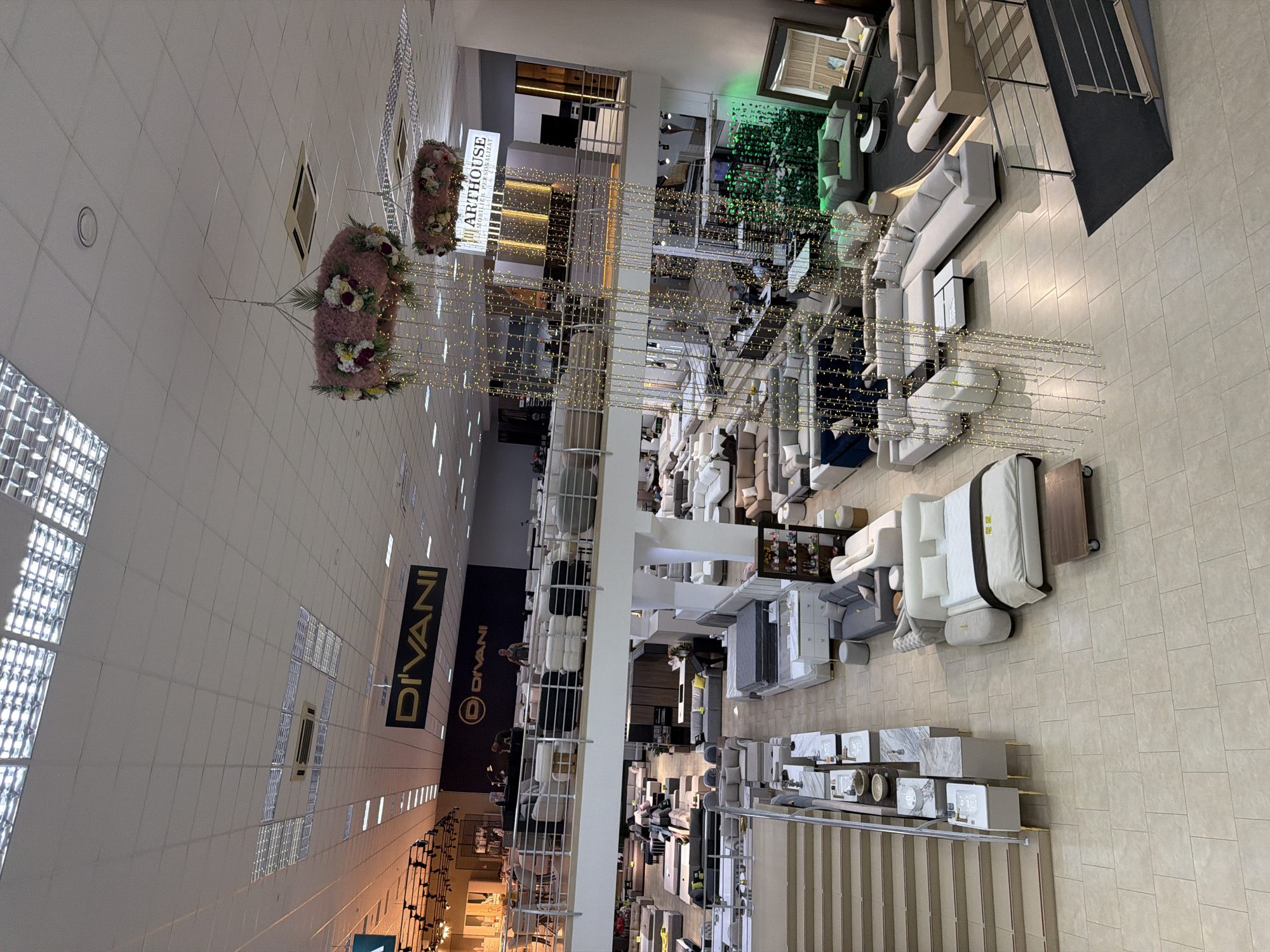 This is a high-angle, wide shot of a large, multi-level furniture store, likely located in Trușeni, Moldova, given the presence of "DIVANI" signage which is a common furniture retailer name in Moldova. The store displays a vast array of sofas, sectionals, beds, dressers, and accent furniture. The store's interior features a modern, open-plan design with multiple levels connected by staircases and walkways. The lighting is bright, coming from fluorescent ceiling fixtures.
On the upper level, a large banner with the text "DIVANI" is prominently displayed. Another banner below it also reads "DIVANI" with a logo. Several people are visible on this level, some appearing to be staff and others customers browsing the merchandise.
The mid-level showcases more furniture, including a variety of sectionals and sofas. Hanging from the ceiling are decorative floral arrangements intertwined with strings of small lights, creating a festive atmosphere.
The ground level is densely populated with furniture displays. On the left, a wide staircase leads down. In the center and right of the frame, numerous sofas, beds, and bedroom sets are arranged for display. Some customers are seen examining the furniture on this level as well.
The store appears to be well-organized, with furniture sections clearly demarcated. The flooring is tiled with large, light-colored tiles throughout. A large framed mirror with ornate detailing is visible on the right side of the ground floor.
