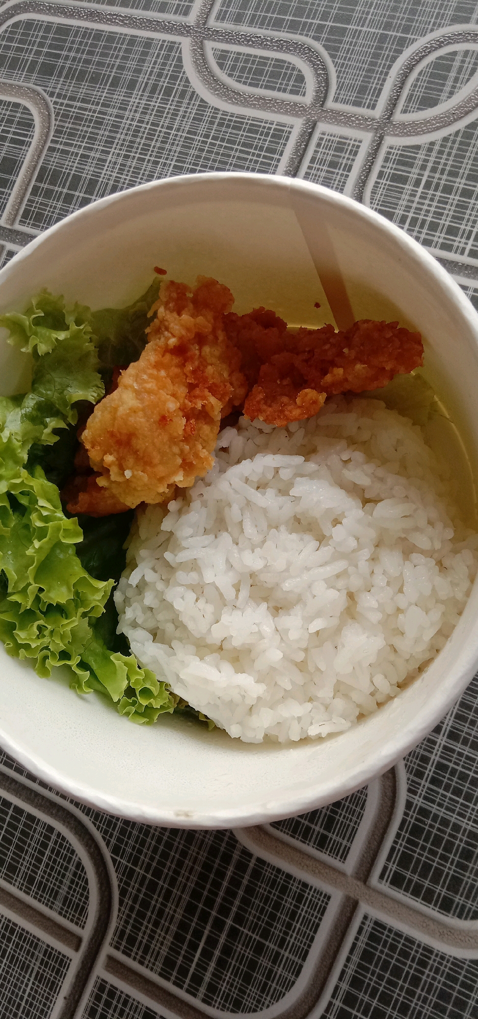 A close-up overhead shot shows a takeout container filled with white rice, two pieces of fried chicken, and a few leaves of lettuce. The container is white and paper. The background is a patterned surface with a dark grey base and a light grey geometric design. The scene is indoors and appears to be a meal on a table. There are no people visible in the image. The meal consists of a portion of white rice, two pieces of crispy fried chicken, and a small amount of fresh green lettuce.