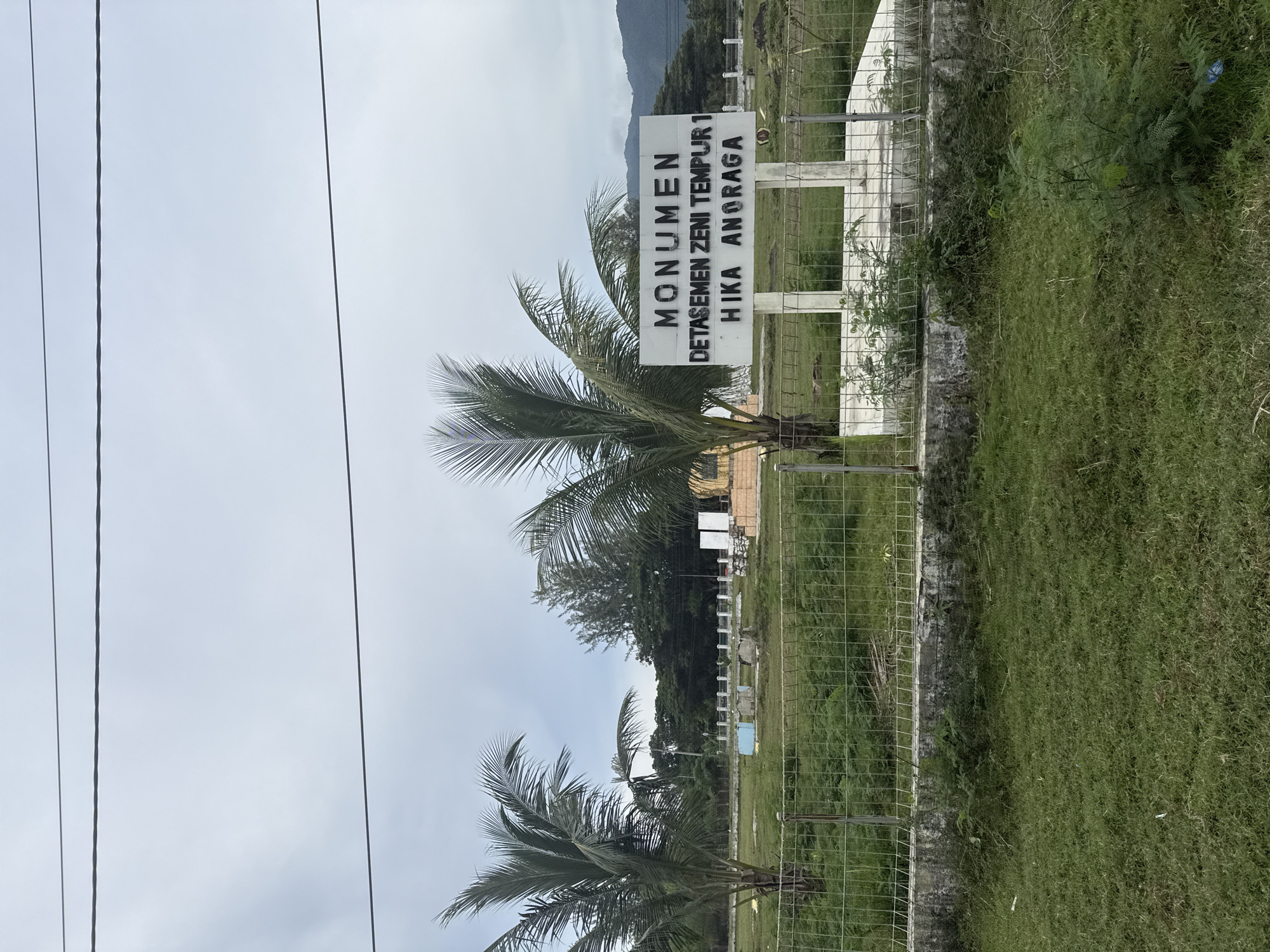 The image depicts an outdoor scene at the city of Lamkruet, Indonesia. In the foreground, a metal fence separates a grassy area from a field of green vegetation. Beyond the fence, a white monument stands with black lettering that reads "MONUMEN DETASEMEN ZENI TEMPUR 1 HIKA ANORAGA". Two palm trees are visible to the left of the monument. In the background, there are more trees and some structures, with hills in the far distance under a cloudy sky. Power lines are visible at the top of the image. The environment appears to be a rural or semi-rural area with a monument and natural vegetation.