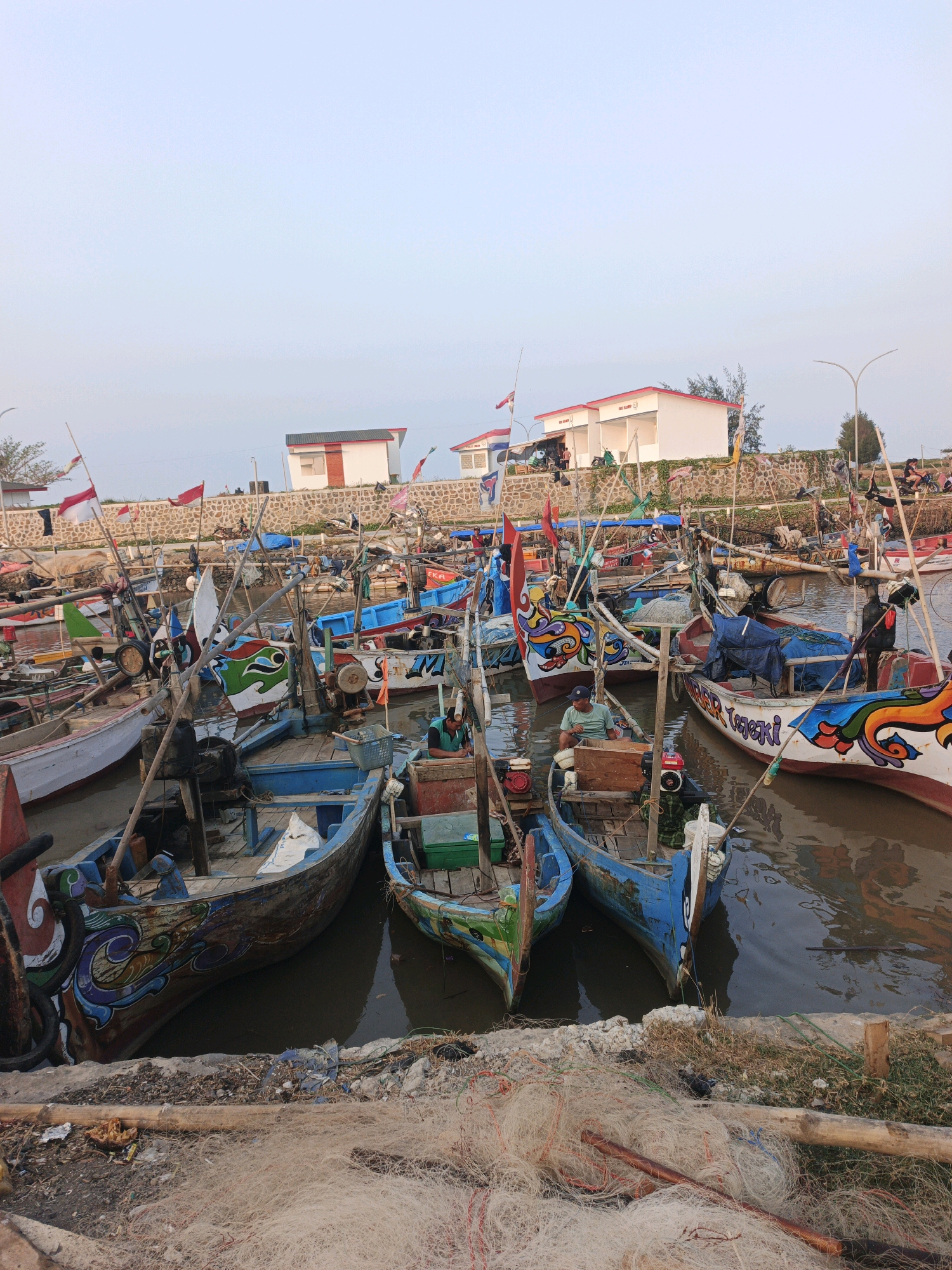The image depicts a bustling harbor scene in Bumiharjo, Indonesia, likely during daylight hours with an overcast sky. Numerous traditional fishing boats, characterized by their colorful, intricate designs and varying sizes, are moored together in the water. Some boats are empty, while others have individuals on board, engaged in activities related to fishing or maintenance. Fishing nets are visible both in the water and piled on the shore in the foreground. In the background, a low, stone wall separates the harbor from a row of modest, white buildings with red roofs. Trees and flagpoles, some adorned with Indonesian flags, are also present. The overall impression is one of a working fishing community.