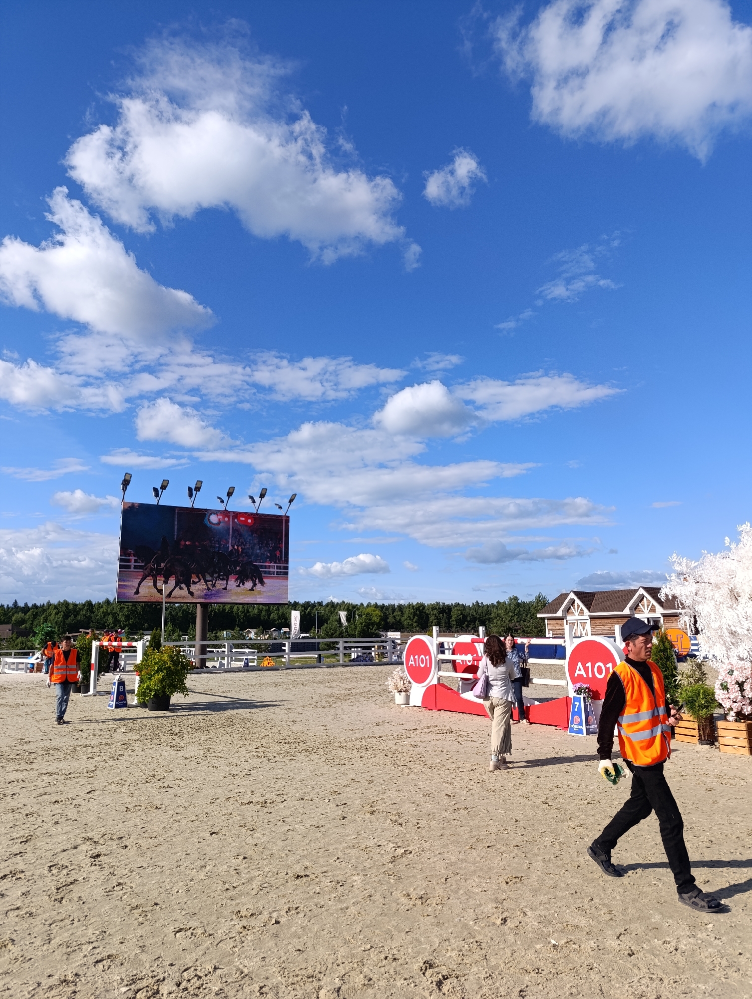 This is a medium-shot, eye-level, outdoor photograph taken on a sunny day with a bright blue sky and scattered white clouds. The scene depicts an equestrian event in a sandy arena.
In the foreground, a man wearing an orange safety vest, dark pants, and a baseball cap is walking to the right. He is holding a mobile phone and what appears to be a small green object in his gloved hands. Further to the left, another person in an orange safety vest is visible in the background, walking towards the left.
The arena itself is covered in sand, with white fencing lining the perimeter. A large LED screen is prominently displayed, showing an image of several horses in motion. Red and white barriers with the text "A101" are set up in the middle ground, with some decorative white floral elements and potted plants. In the far background, there is a line of trees and some buildings, including a wooden structure with gabled roofs.
The lighting suggests it is daytime, likely late morning or afternoon, given the clear sky and the shadows cast. The overall atmosphere appears to be one of an organized event, possibly a competition or show, with staff present to manage the area. The image is clear, capturing the details of the sandy ground, the signage, and the people engaged in their roles at the event.