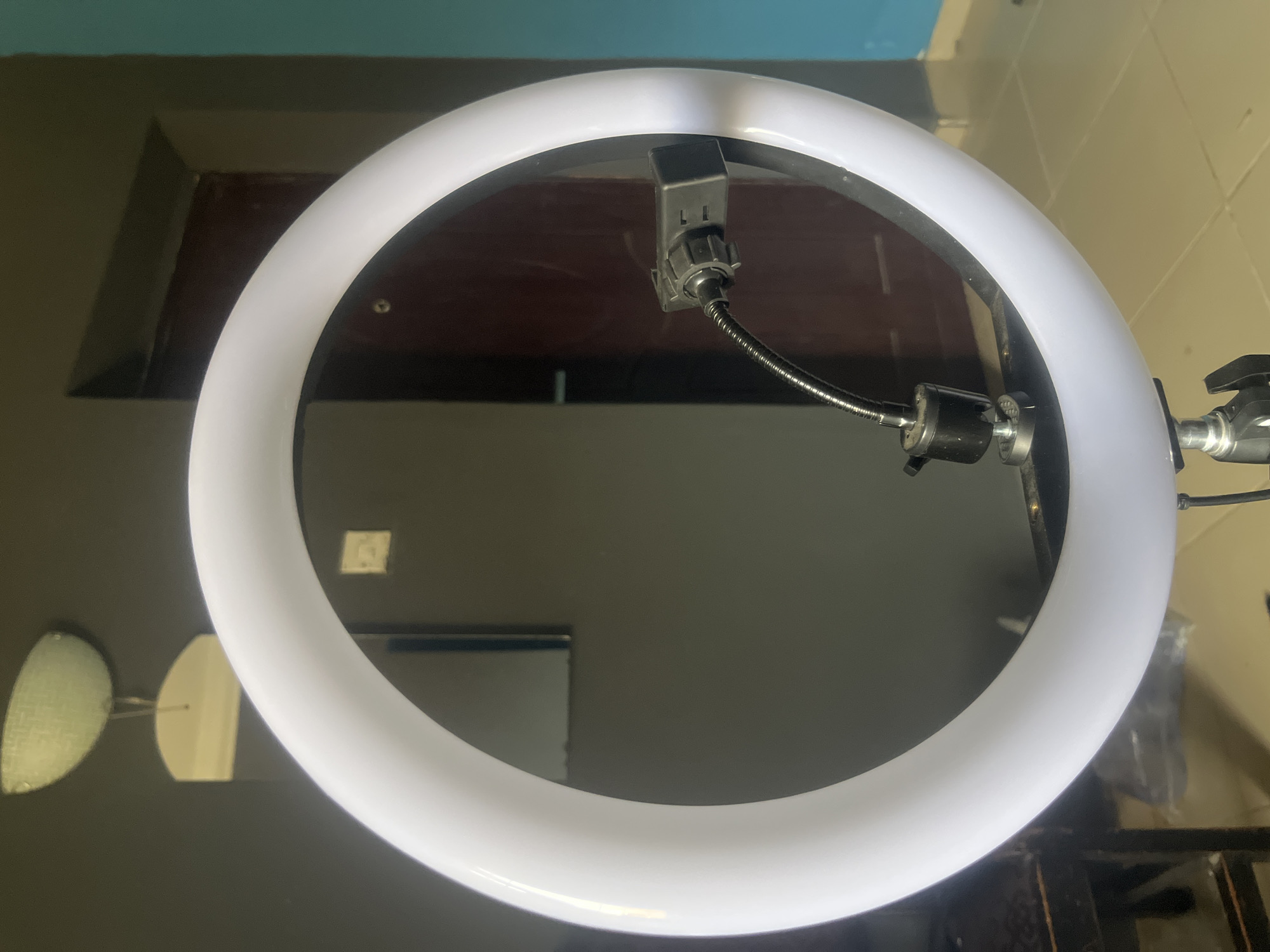 The image shows a close-up of a circular LED light, commonly used for photography or videography. The light is white and illuminated, creating a bright halo. Attached to the light is a black mounting bracket with a flexible gooseneck arm extending downwards, which likely holds a phone or camera. The background is dimly lit and appears to be an indoor setting. A dark brown wooden surface is visible behind the light, and a wall painted in muted tones of grey and turquoise is present. Reflections can be seen on the dark surface, including a blurry image of a light fixture with oval shades. The overall impression is that of a staged setup for content creation, possibly in a home or studio environment. The time of day and weather are not discernible, nor are any specific emotions. No visible text is present in the image. The setting is given as Barakin Akawo, Nigeria.
