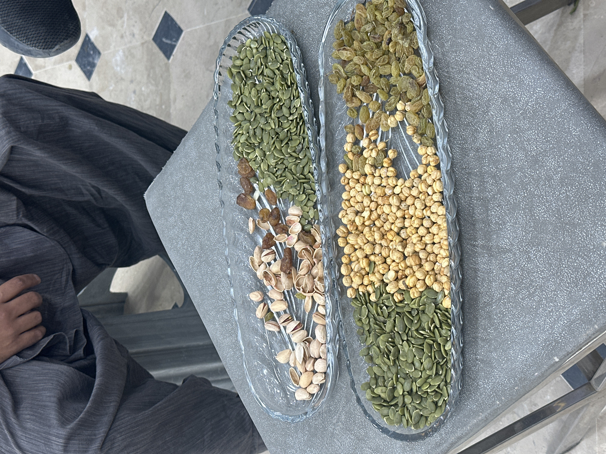 The image shows two glass trays containing snacks, likely in Quetta City Tehsil, Pakistan. The tray on the left has green pumpkin seeds, a few brown raisins, and pistachios. The tray on the right has green raisins, small beige chickpeas, and green pumpkin seeds. The trays are placed on a gray surface. The left side of the image shows a person seated, wearing a gray outfit, with a partial view of their hand. The floor has a diamond pattern.