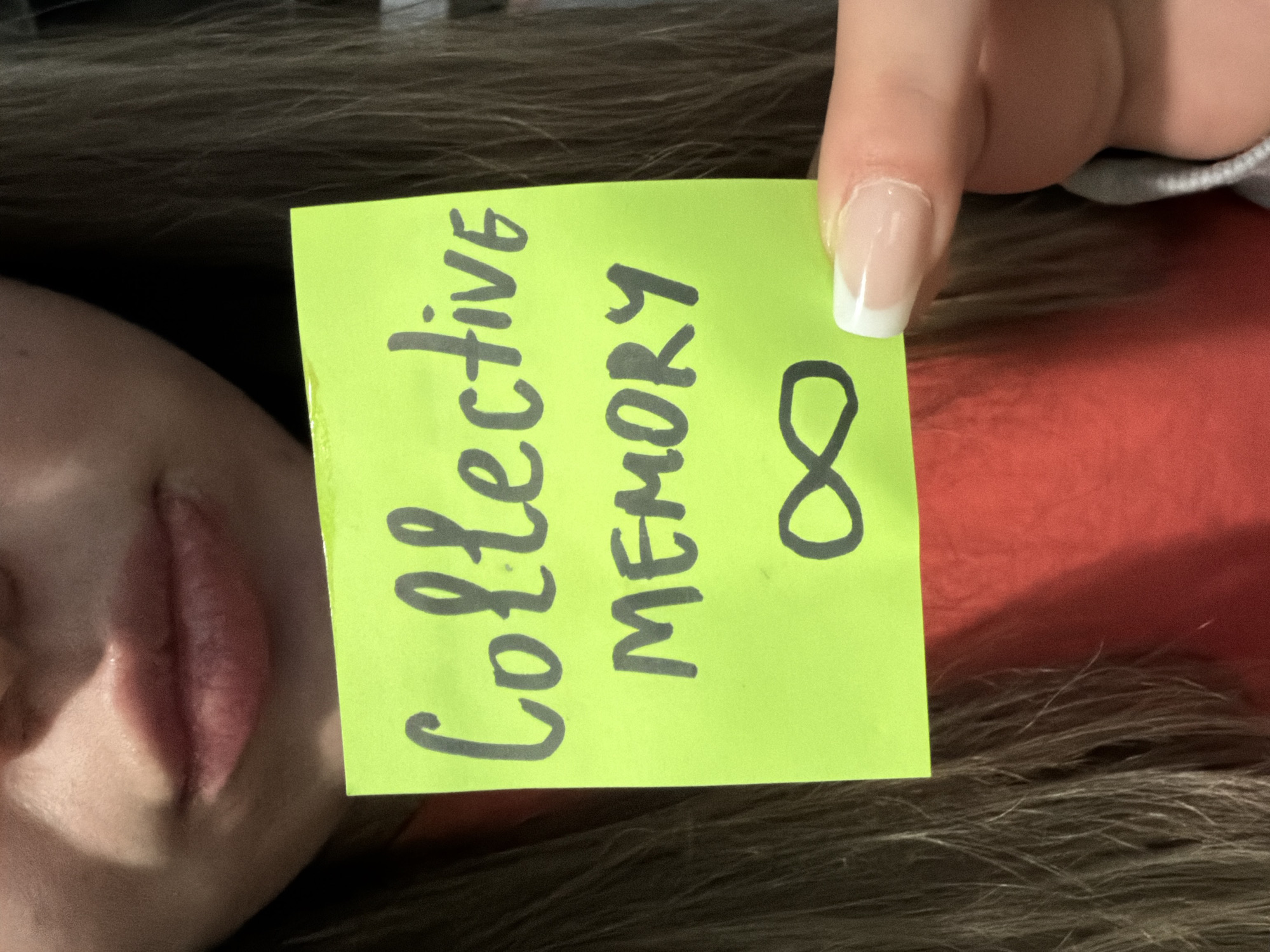 The image shows a person holding a bright green sticky note with the words "Collective MEMORY ∞" written in black marker. The person's face, specifically the lips and part of the cheek, is visible to the left of the sticky note. A hand with a manicured nail is holding the top right corner of the note. The background includes brown hair, a grey fabric and a red surface. There is no indication of the city of Varna, Bulgaria.