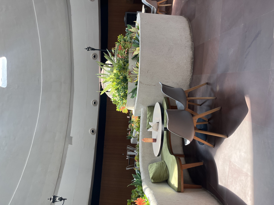 The image shows a seating area in an indoor space located in Sahibzada Ajit Singh Nagar, India. The space is characterized by a white, rounded ceiling with a central skylight, and beige, tiled flooring. There are two white, rounded planters filled with greenery, and two gray, rounded planters containing plants. There are two black lamp posts and two white, round, light fixtures. A circular table with a wood top and metal base sits between two gray chairs with wood legs. A curved, green bench sits against a white wall, and a white, rounded planter with greenery rests behind the seating area. The scene is empty with no people present.