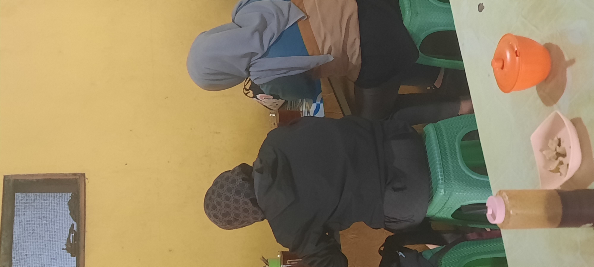 In Selopuro, Indonesia, two individuals are seated at a table with a yellow wall as the backdrop. The individual on the left wears a dark hooded jacket and a patterned headscarf. The person on the right is dressed in a blue and tan top with a light blue headscarf. Both are seated on green plastic stools. On the table in the foreground, there is a brown bottle with a pink cap, a small white dish containing white food items, and an orange lidded container casting a shadow. A small window with a screen is visible in the upper left corner of the image.