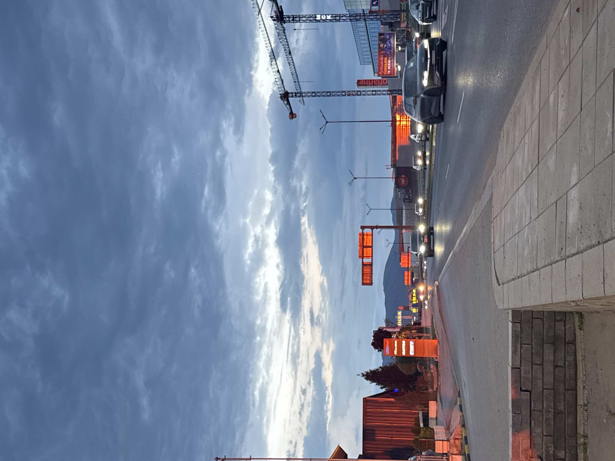 The image is an eye-level, outdoor shot taken on a sidewalk along a busy street. The scene is likely in Sofia, Bulgaria, based on the presence of IKEA and other European retail signs. The camera is oriented at an angle to the street, showing the curb and sidewalk on the right. Vehicles, including cars with headlights on, are traveling along the street, creating streaks of light.
In the background are buildings, construction cranes, street lights, and illuminated signs for various businesses. The sky is filled with clouds, suggesting either dawn or dusk, and the lighting is dim but supplemented by artificial light sources. The street is wet, possibly from recent rain, reflecting some of the light. The overall impression is a typical urban evening scene with active traffic and commercial activity.