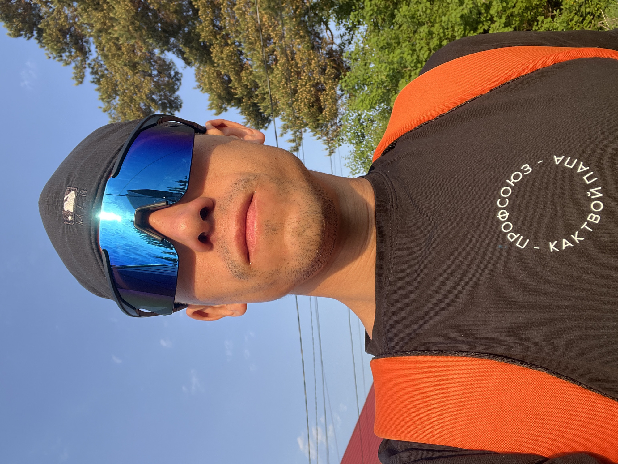 A man is seen in an outdoor setting in Chaikovsky, Russia. He is wearing a black cap with a baseball logo, sunglasses with reflective blue lenses, and a black shirt with an orange trim around the shoulders and a white circular design with Cyrillic text. The background includes a blue sky, a tree with green leaves, and potentially part of a building or structure. The photograph appears to be taken during daylight hours.