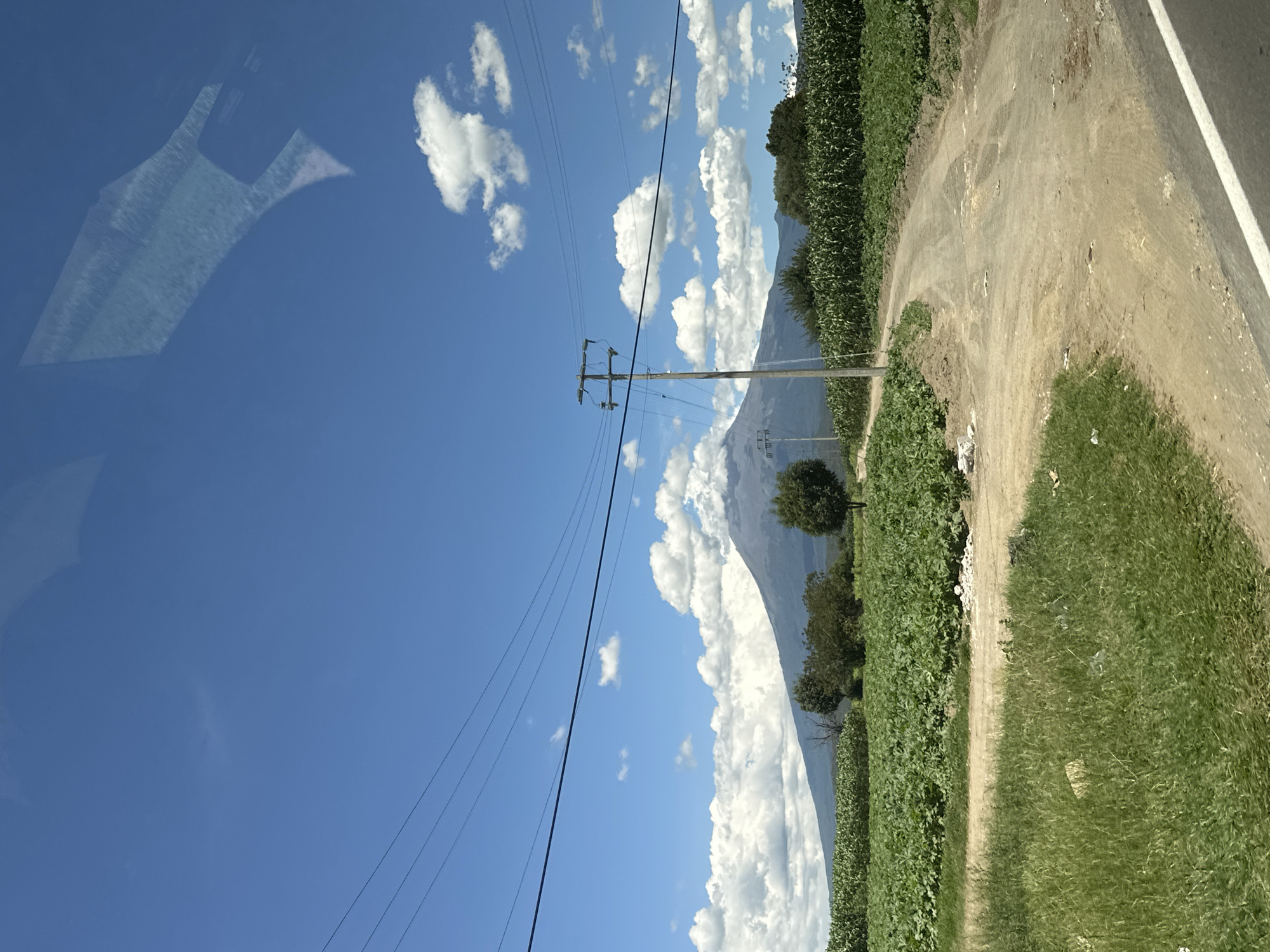 The image depicts a rural landscape under a bright blue sky with scattered white clouds. A dirt road curves through a field of green crops, with a power line stretching across the frame. In the distance, a large, hazy mountain dominates the horizon. The foreground shows lush green grass and some scattered debris along the roadside. The scene appears to be in the vicinity of San Miguel Papaxtla, Mexico, a region known for its agricultural land and prominent geological features.