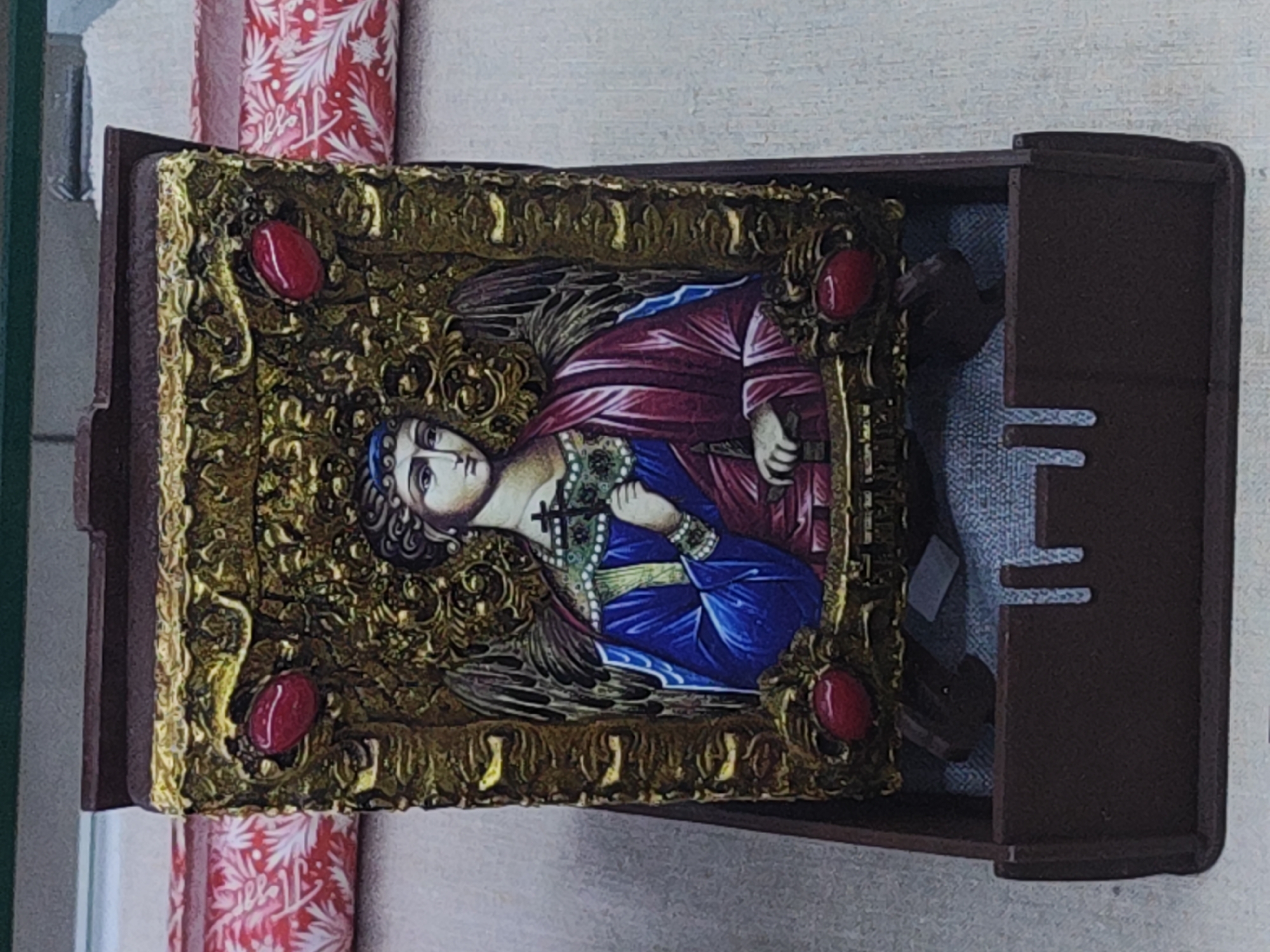 An ornate religious icon is displayed upright within an open dark brown wooden box, resting on a light-colored, possibly textured, surface. The icon depicts a haloed figure, likely an angel or saint, rendered in a traditional Eastern Orthodox style. The figure has fair skin, dark hair, and wears a blue headpiece. Its richly colored robes are deep red on the viewer's right side and vibrant blue on the left, with visible wings extending from its back. In its right hand, the figure holds a staff-like object, and in its left, a small, dark cross. The serene face looks slightly upwards and to the viewer's left. The icon is encased in an intricate golden frame adorned with raised, scrolling patterns and accented by four symmetrically placed oval, deep red cabochon jewels.
The dark brown wooden box, with its lid fully open, reveals a light gray, textured fabric lining inside. Horizontal notches are visible on the inner right wall of the box. Behind the box, a cylindrical object wrapped in festive red and white patterned paper is partially visible. The paper features a stylized white foliage pattern and discernible cursive text that appears to read "Hello." The scene suggests an indoor display, possibly in a shop or home in Saint Petersburg, Russia.