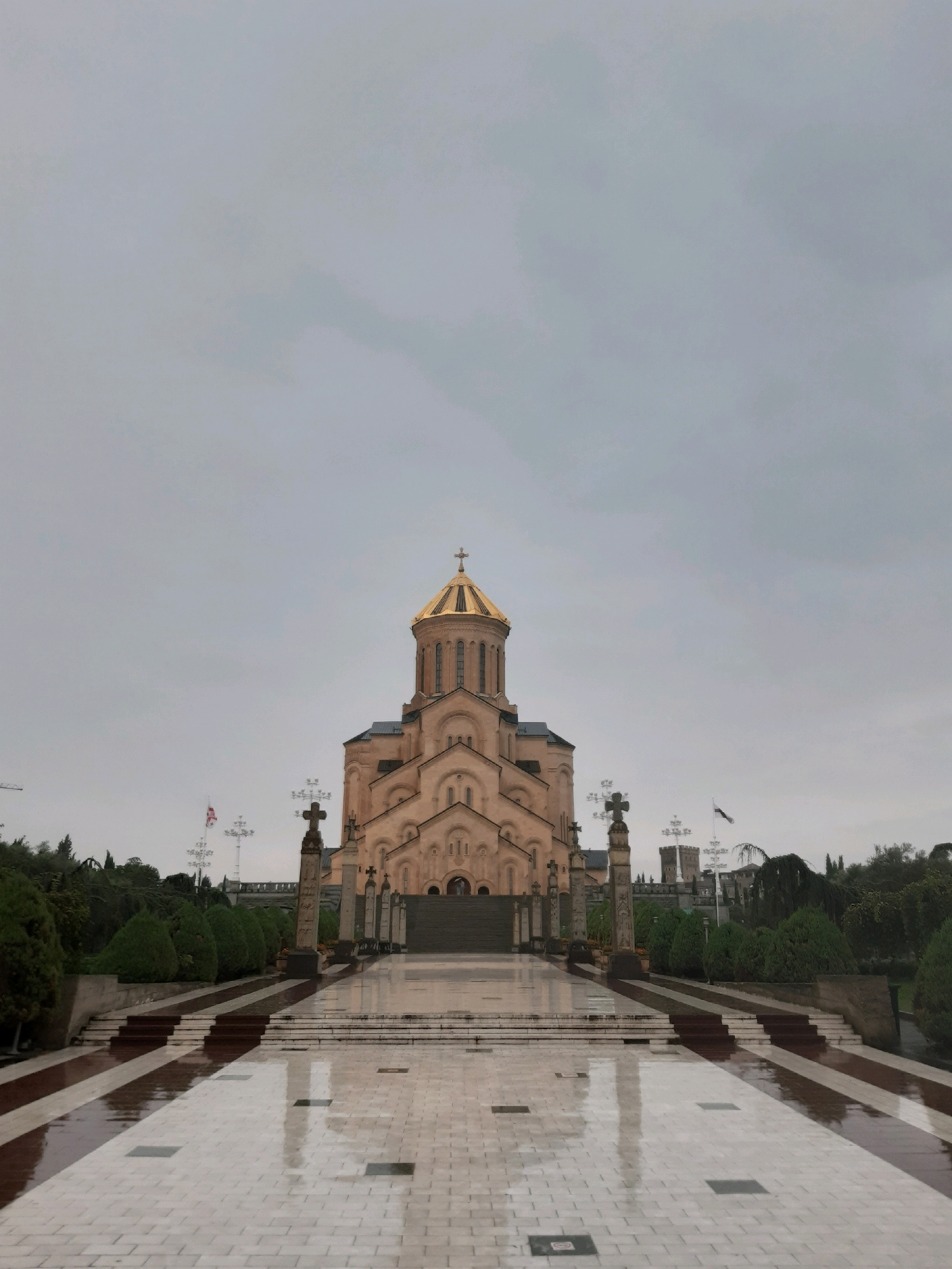 The image shows the Holy Trinity Cathedral of Tbilisi, Georgia, under an overcast sky. The cathedral is a large, prominent religious structure with a golden dome, made of beige stone. It is situated at the end of a wide, wet, paved walkway that reflects the surroundings. The walkway is lined with manicured green bushes and trees. Several tall crosses stand on either side of the walkway, leading up to the cathedral's entrance. A Georgian flag flies on a flagpole to the left of the cathedral. The ground appears wet, suggesting recent rain. The scene is symmetrical and grand, with the cathedral as the central focus.