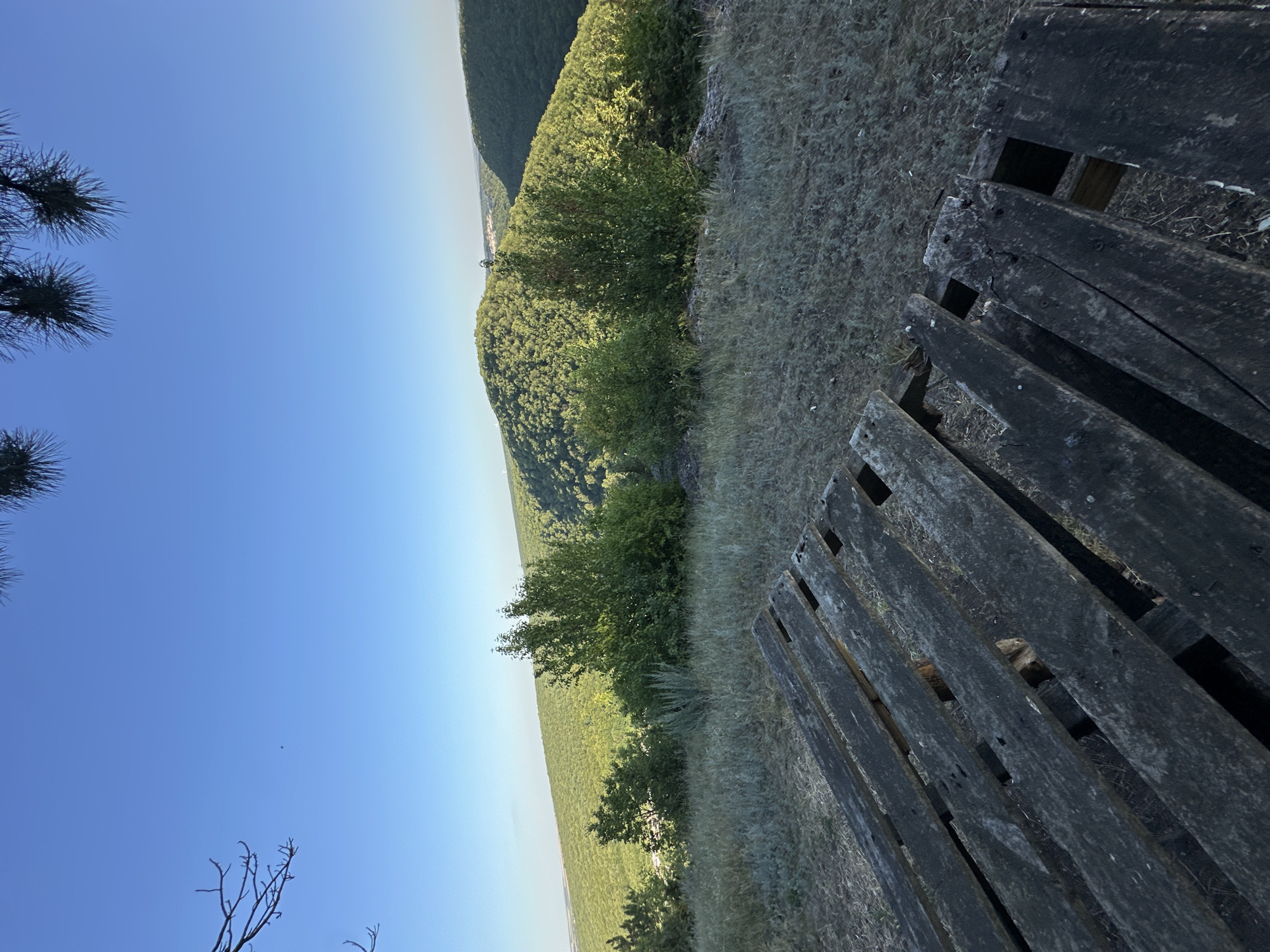 The image depicts a scenic overlook in the city of Pryvorottia Druhe, Ukraine. In the foreground, a weathered wooden structure, possibly a picnic table or bench, is positioned on dry, grassy terrain. The structure is composed of rough, dark wooden planks with visible gaps and wear. Beyond the foreground, a lush, green landscape unfolds with rolling hills densely covered in trees. The sky is a clear, bright blue with the sun casting gentle light on the scene. The overall impression is one of natural beauty and tranquility.