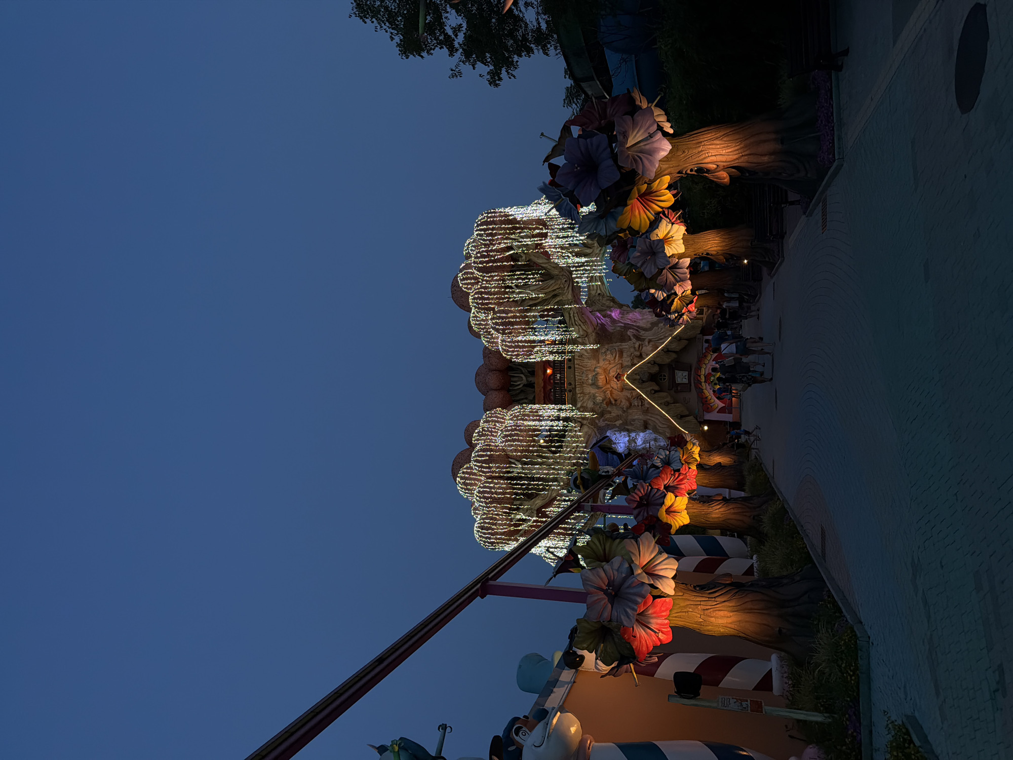 This image captures a whimsical and vibrantly lit scene at dusk, likely within an amusement park or entertainment venue, given the decorative elements and presence of people. The primary subjects are exaggerated, stylized trees adorned with large, colorful artificial flowers in shades of purple, yellow, red, and blue. These trees are wrapped in numerous strands of warm white lights, creating a magical and festive atmosphere.
The scene appears to be a pathway or promenade, with a paved surface on the right side of the frame, suggesting an outdoor public space. Several people are visible, walking along the path and interacting with the environment, though their faces are not clearly discernible. The overall mood is cheerful and inviting, enhanced by the warm lighting and playful design.
Notable details include the time of day, which is twilight or early evening, indicated by the deep blue of the sky. The weather appears to be clear. The architectural style of the illuminated structures and the ground paving suggest a European location, with the provided context identifying it as Castelnuovo del Garda, Italy. The elements strongly suggest a fantasy or fairytale theme. No visible text is discernible in the image.